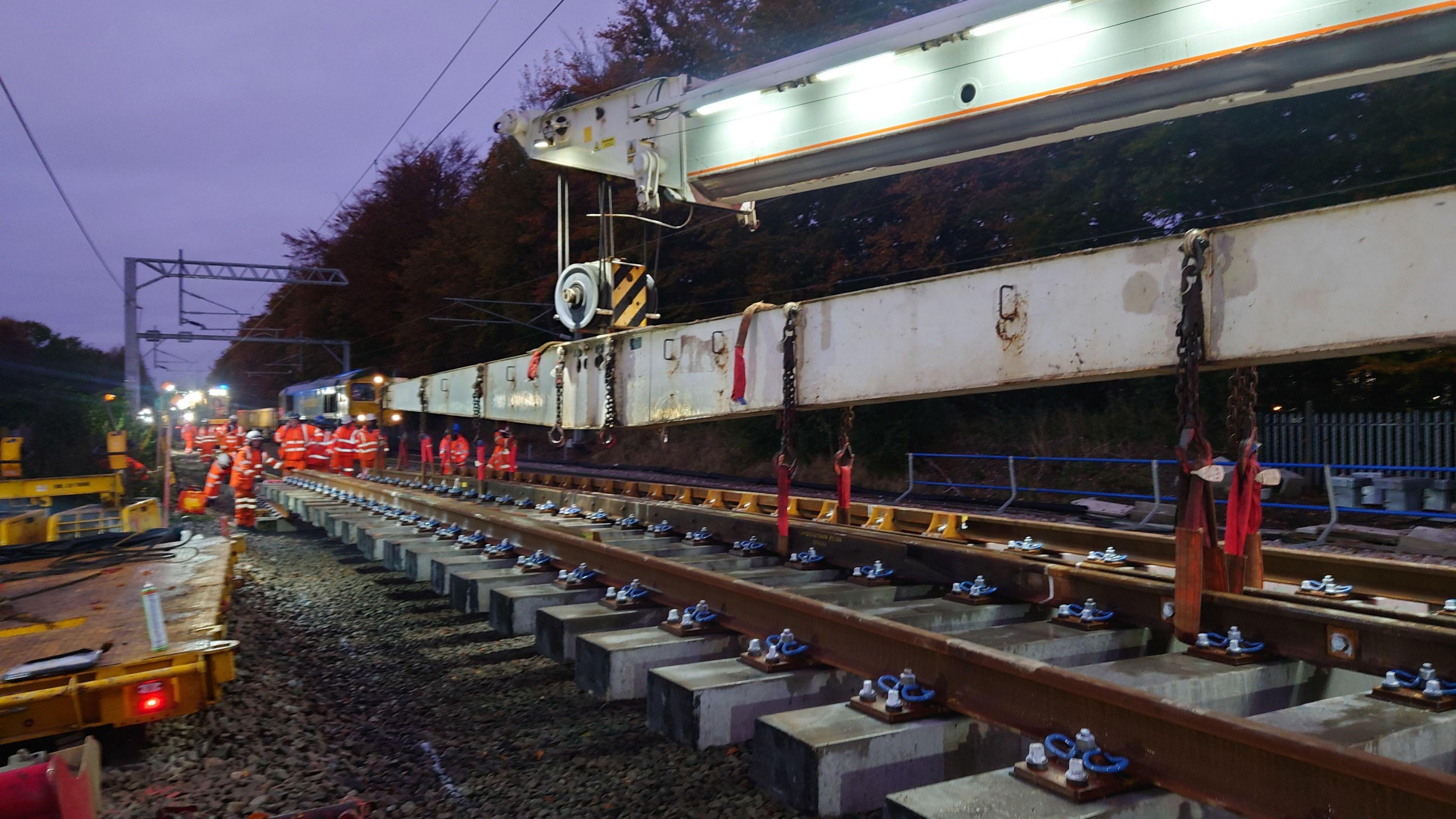 Construction workers wearing orange hi-vis are working on a railway track at night.
