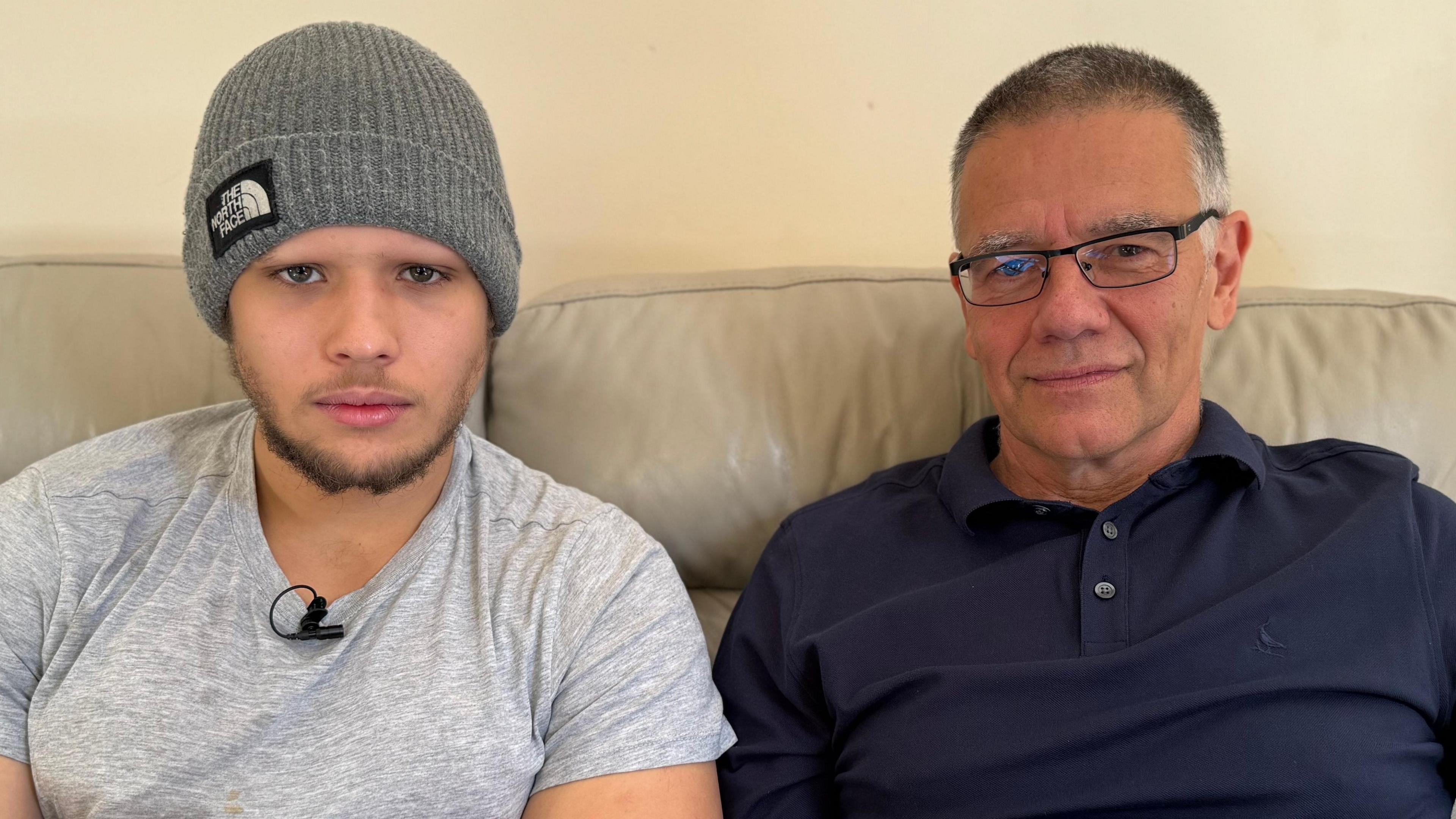 In a grey t-shirt and beanie hat, George sits on a beige leather sofa next to his dad Tom who is wearing a navy top and glasses. Father and son are looking directly at the camera.