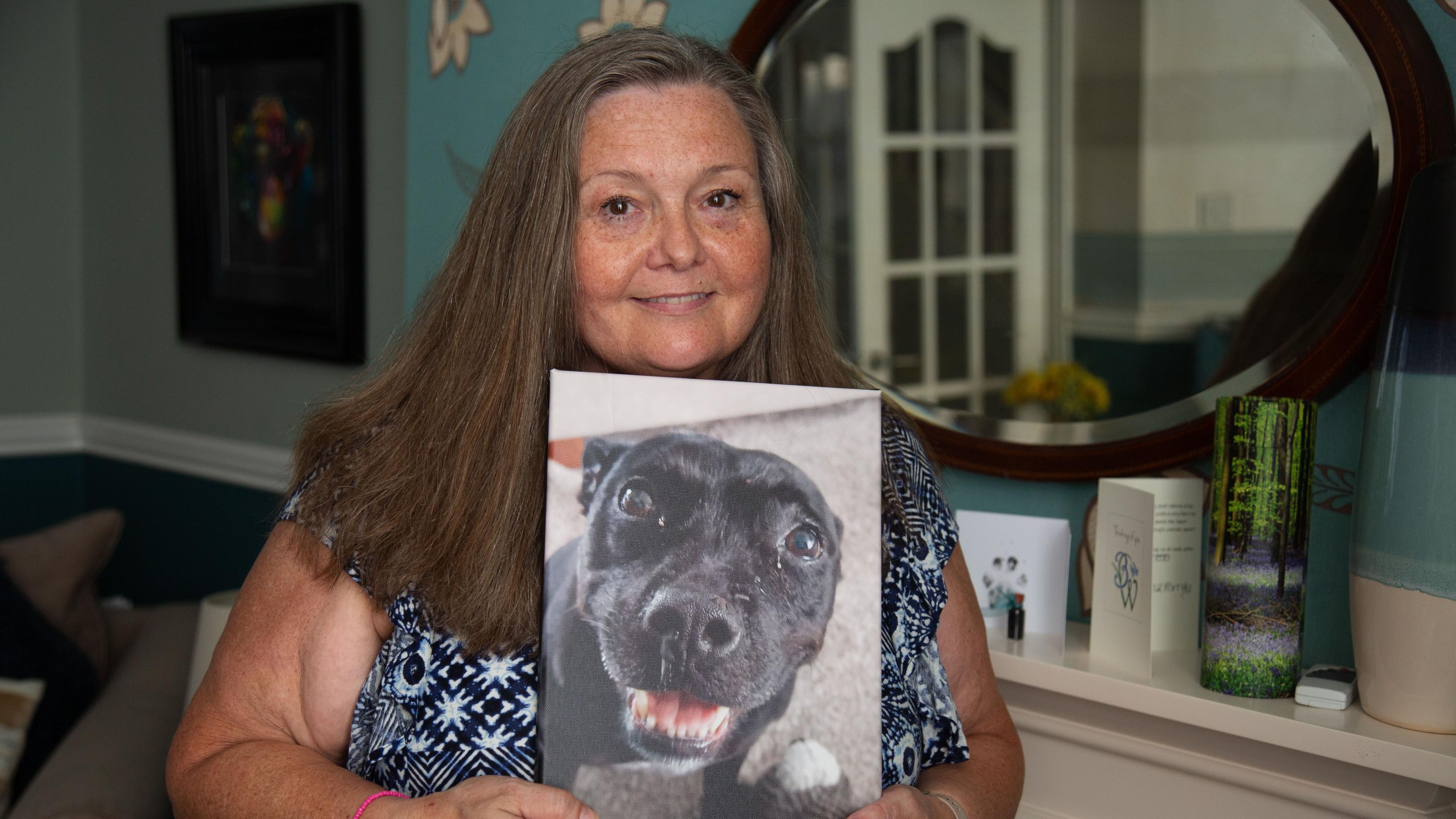 Fiona stands indoors holding a printed photograph of a black dog close to her chest. The print shows a close-up of the dog’s face, with one eye prominently visible and the mouth slightly open. Fiona has long hair and is wearing a patterned top, along with a pink wristband on one wrist.
In the background, a softly lit room is visible with teal-coloured walls, a round mirror mounted on the wall, and a white shelf displaying a card with a paw print and small decorative items, including a tall, green-toned glass ornament.
