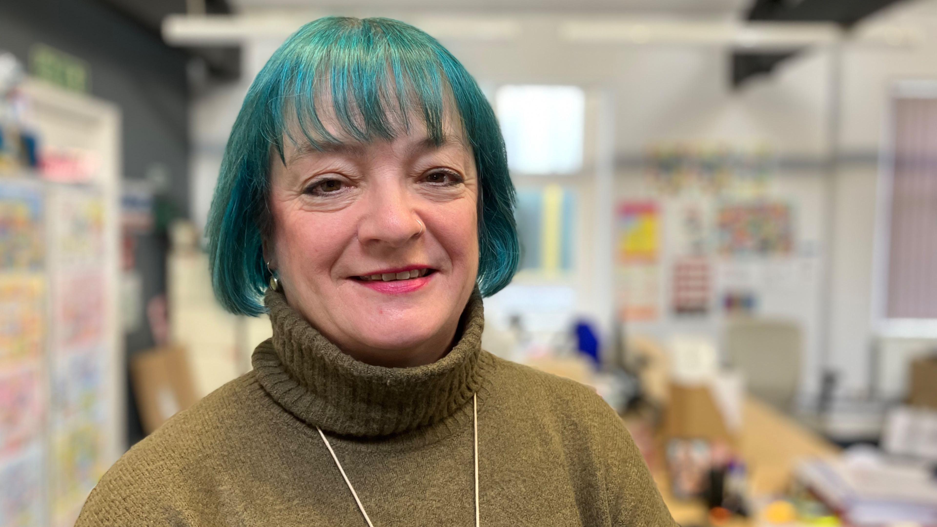 Gill Bainbridge looks into the camera, smiling. She is wearing a yellow-green turtle neck jumper and has a short bob, dyed blue.