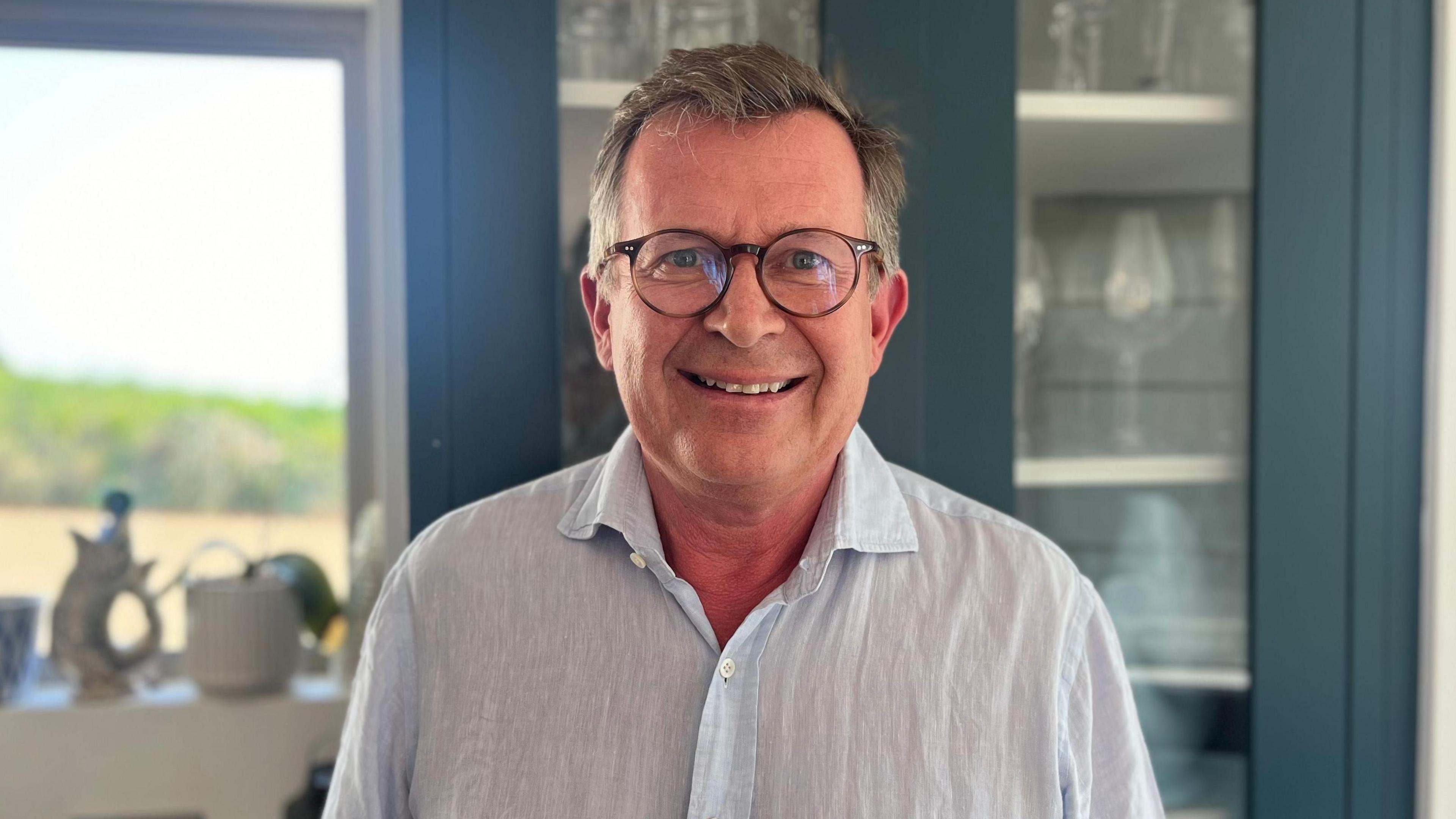 A man wearing black circle shaped glasses and a light blue shirt smiling at the camera in front of a blue cupboard with wine glasses and plates inside it.