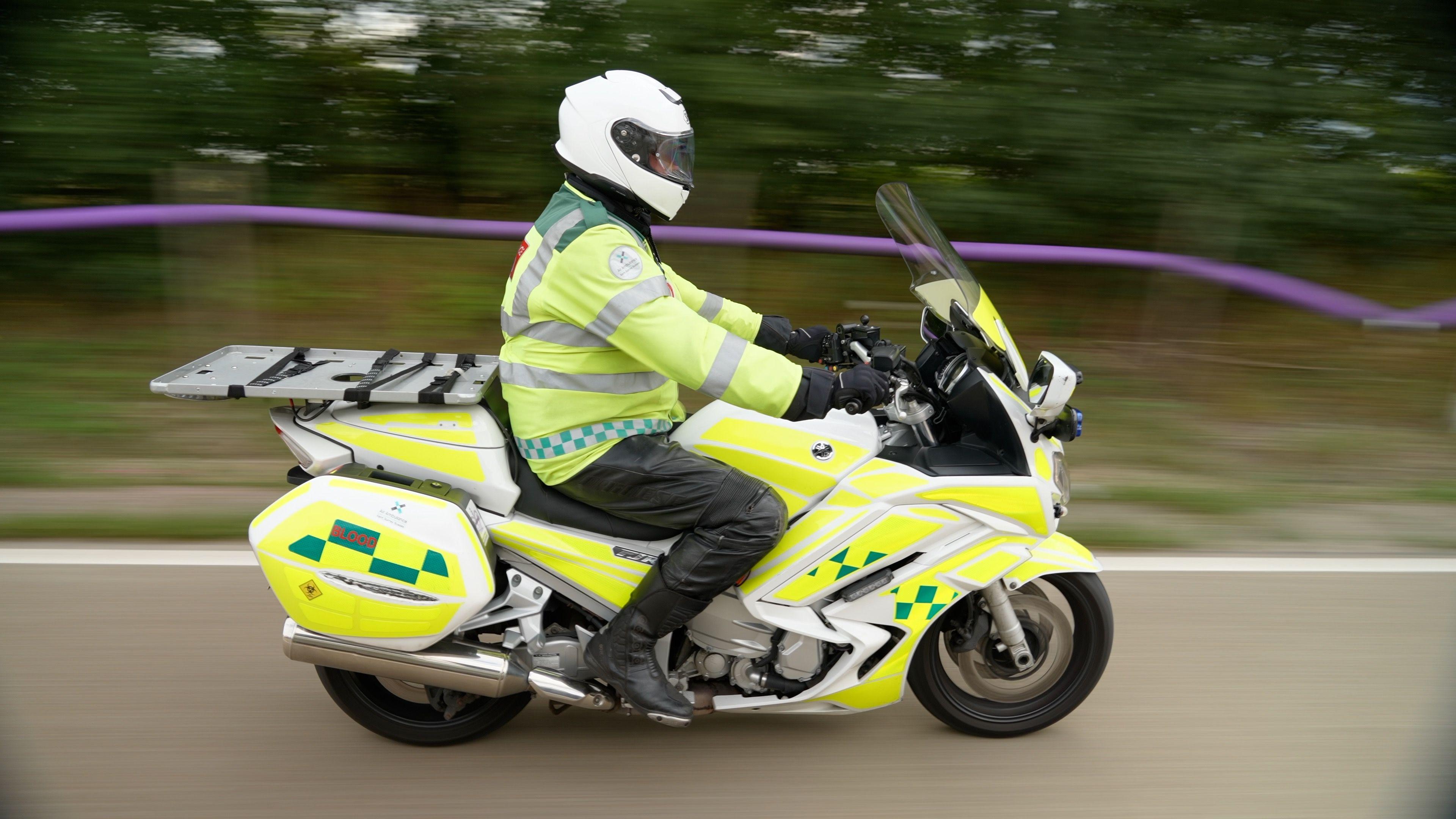 A man wearing a helmet and reflective clothing, driving on a motorbike on the motorway. He is on an ambulance motorbike, which says blood on the side.