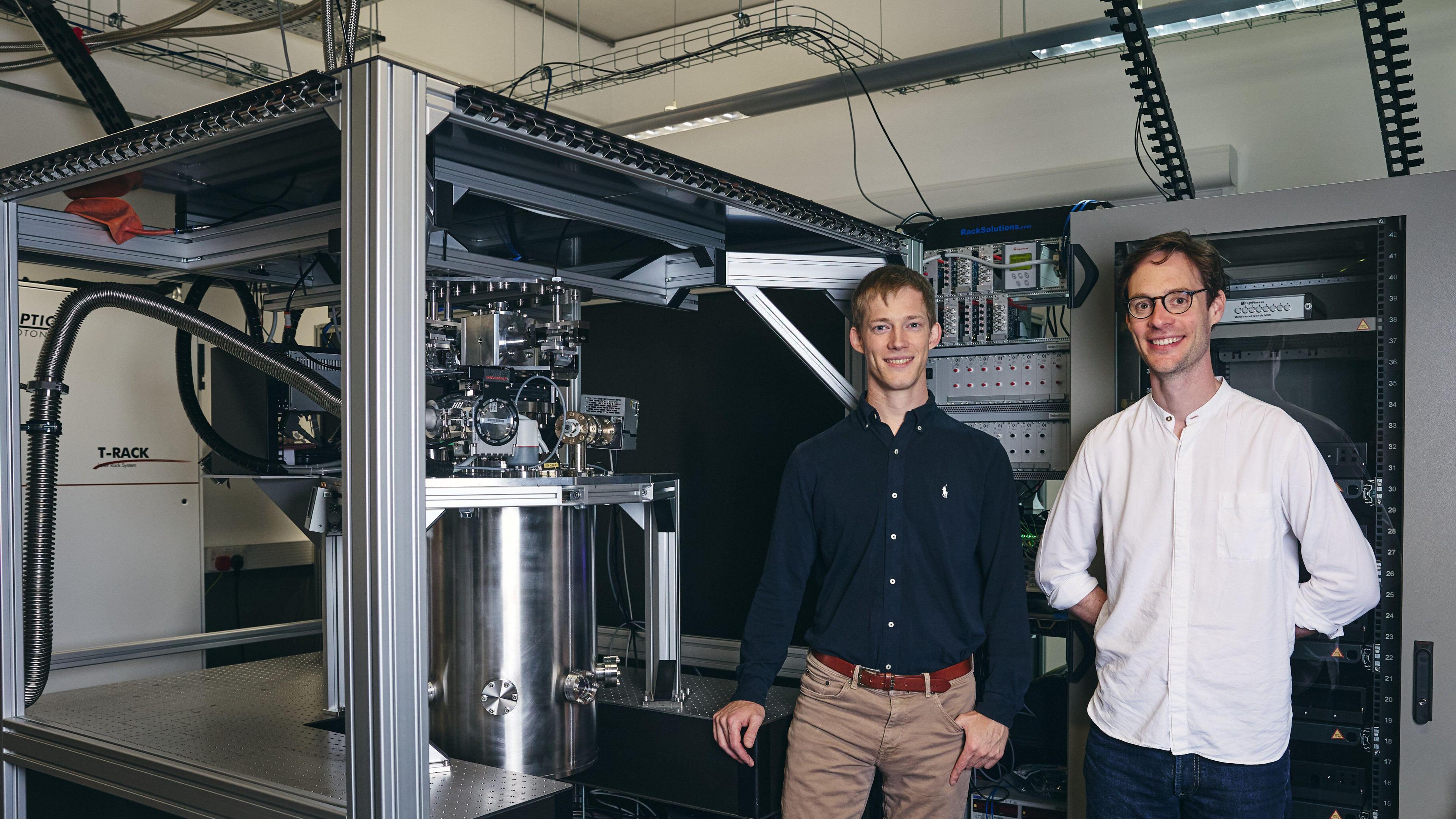 Two men stood in front of a science lab with a quantum chip in. One is fair-haired wearing a blue shirt, whilst the other has dark hair a white shirt and glasses