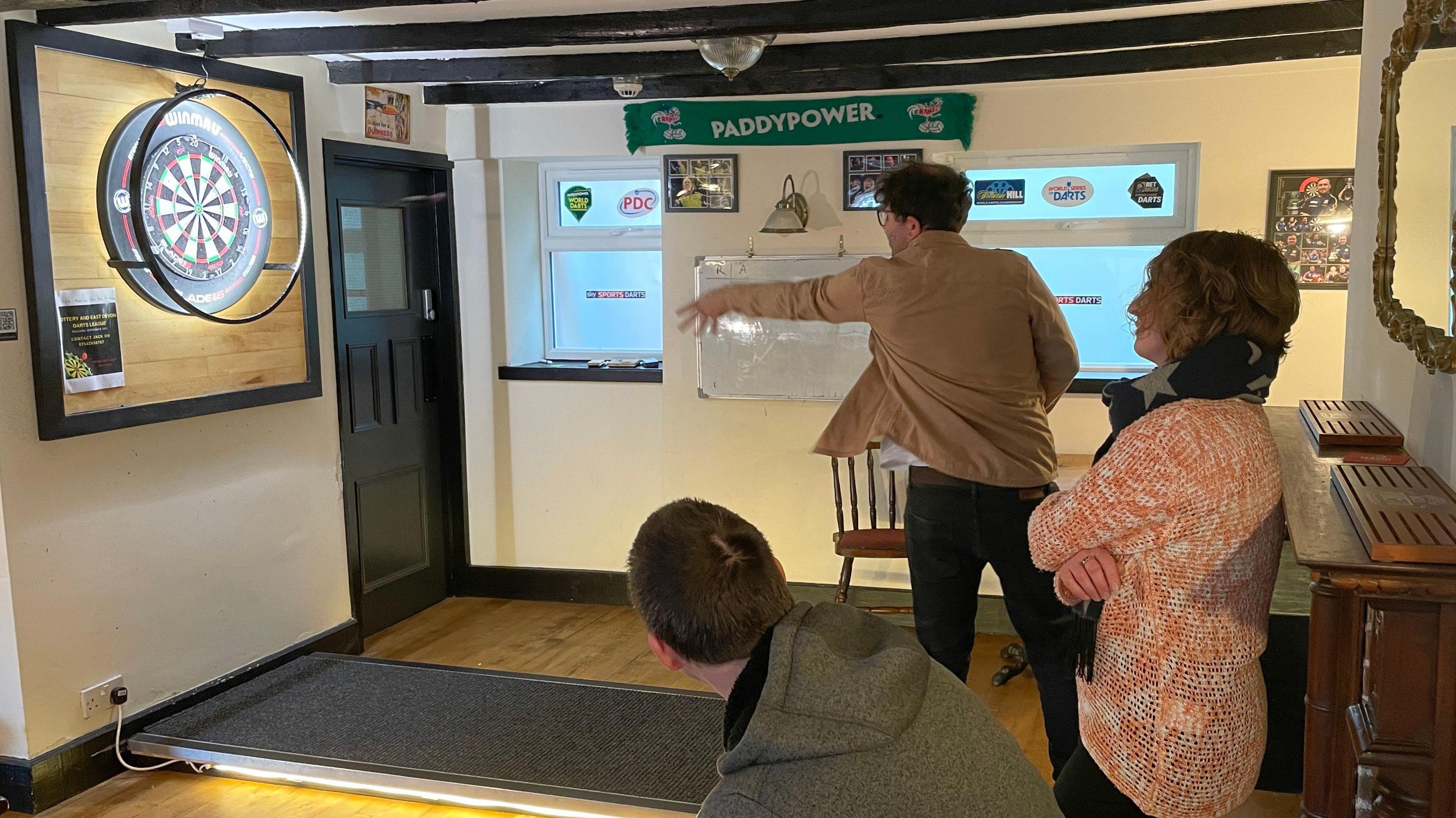 Dan throws a dart at the board with his left hand. His enabler Rhea and his friend Alex watch on.