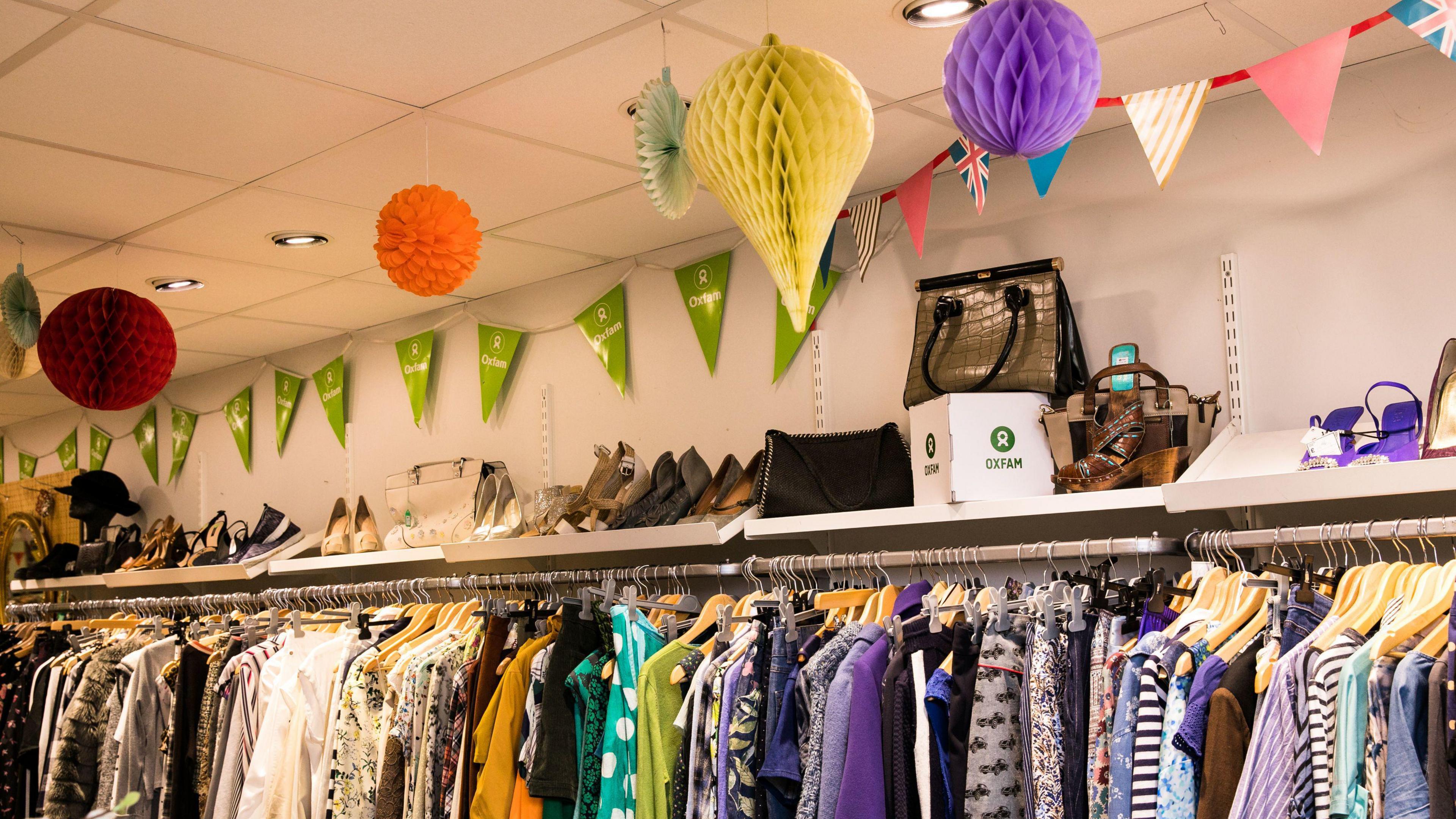 A display in an Oxfam shop in Brighton and Hove. Oxfam-branded green bunting is hanging from the ceiling, along with paper arrangements of different colours. Shoes and bags are displayed on a shelf. Underneath, a rack is packed with various clothes.