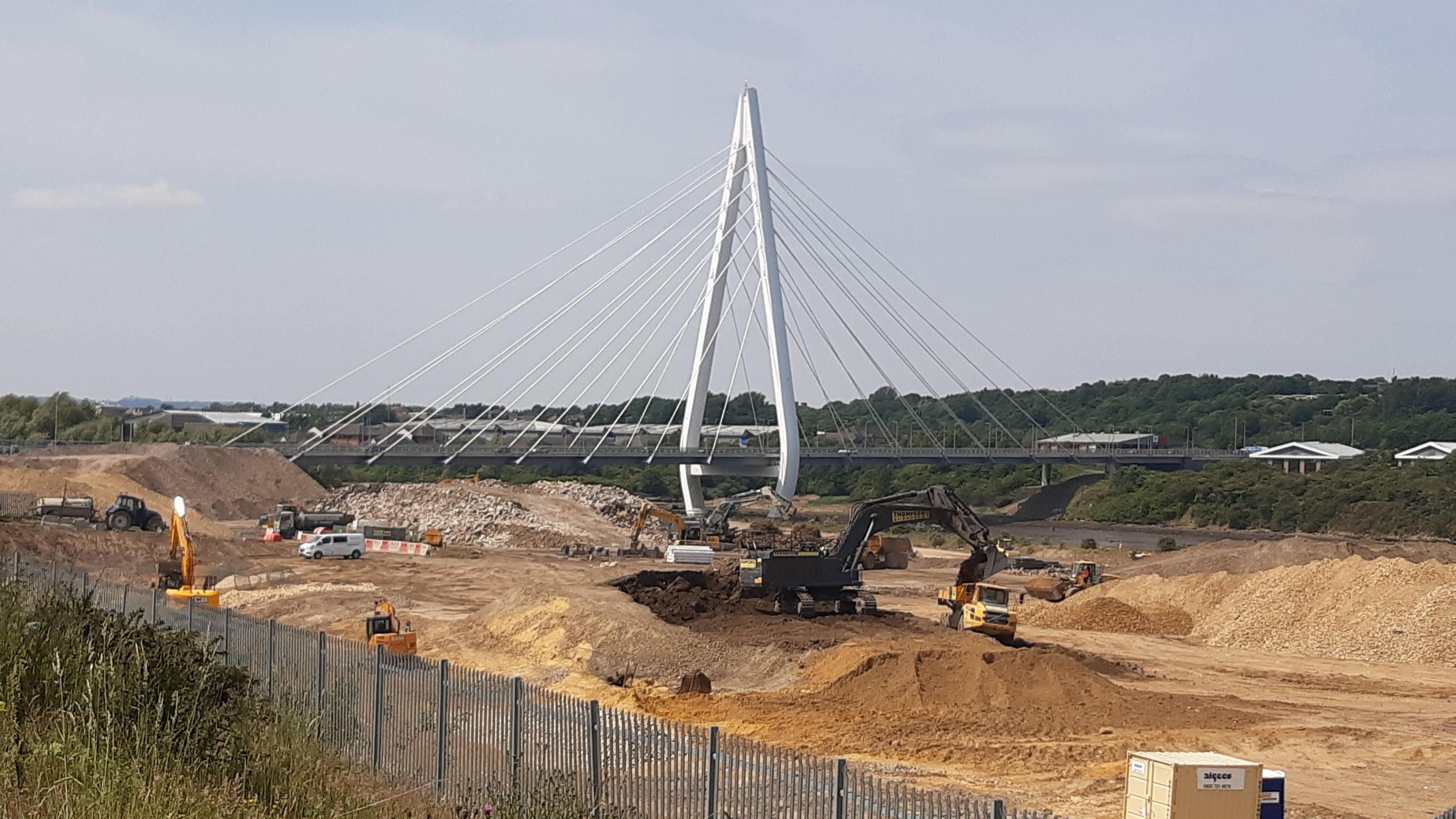 The construction site of the Crown Works film studio. A large area of land is dug out. There are diggers dotted around the site. A white bridge borders is on one side of the site.