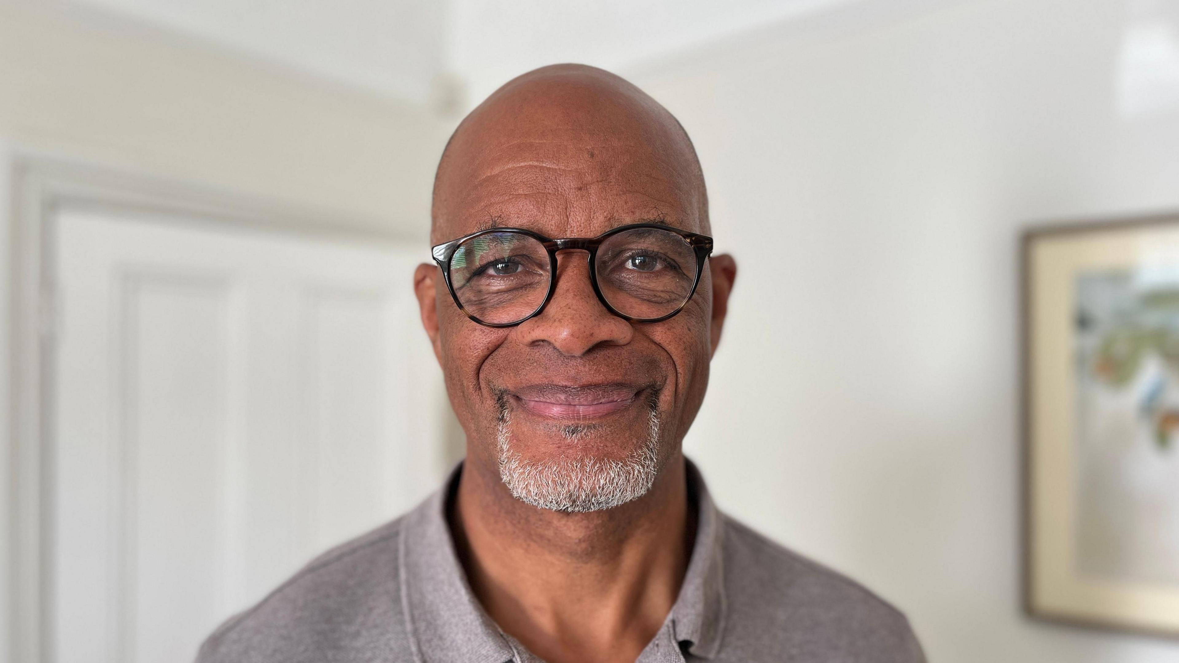 John Lynch is a retired headteacher. He is black with a bald head and wears black-rimmed glasses. He wears a grey polo shirt. He is smiling at the camera.