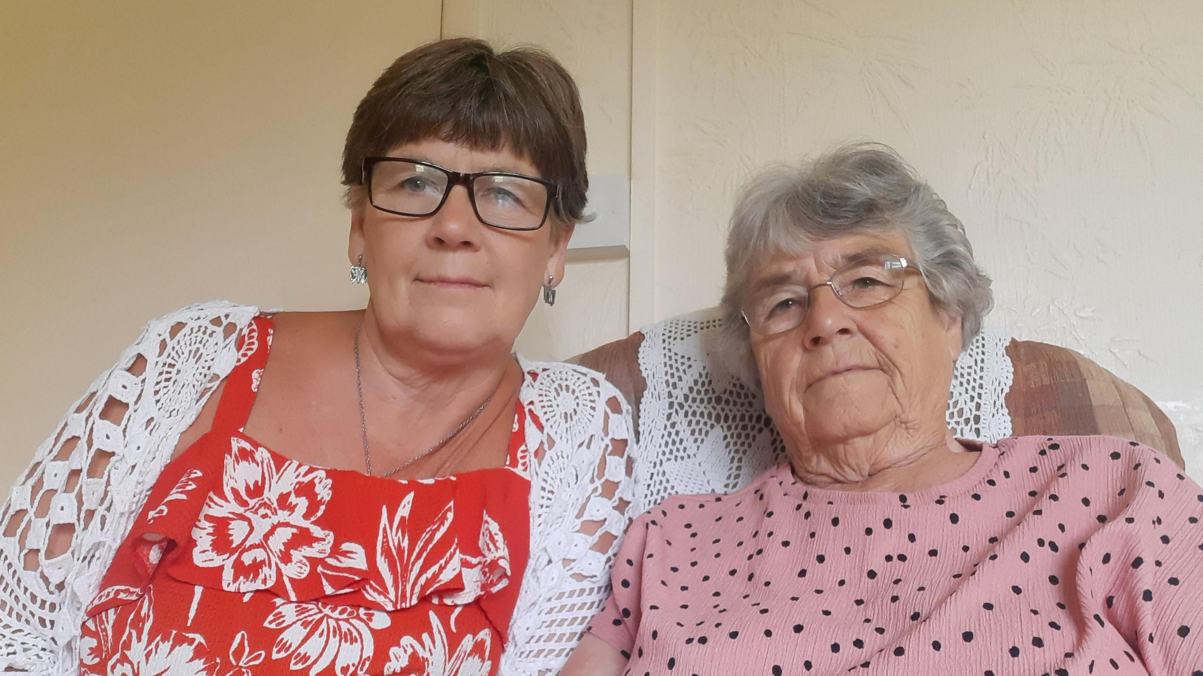 Karen Williamson is wearing glasses and a red flowery top beneath a light white cardigan. Jean Hewitt, who also wears glasses, has a pink, black spotted jumper on. They're sitting together on an armchair.