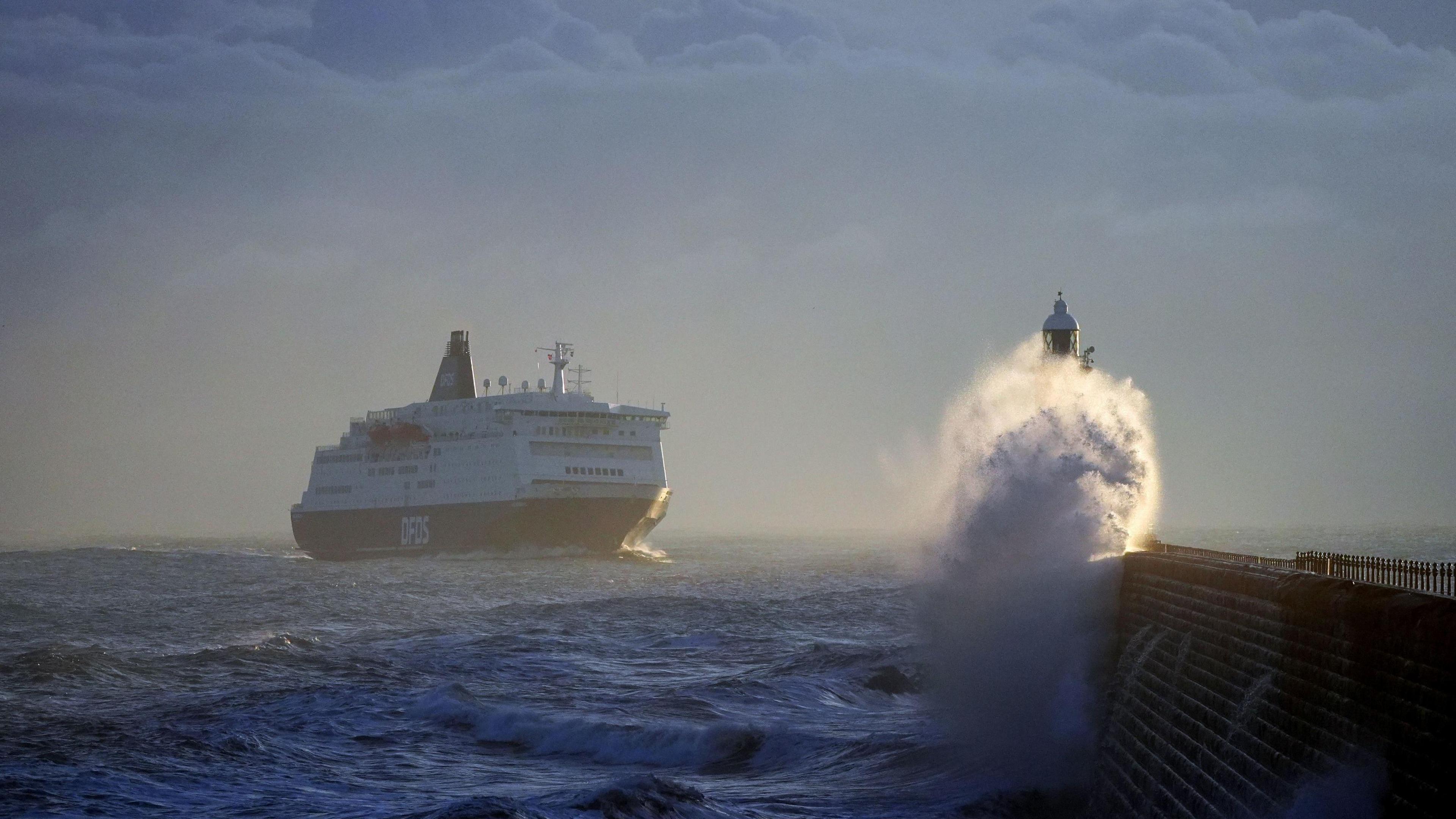 DFDS ferry at sea approaching land where a wave can be seen crashing over a small lighthouse. 