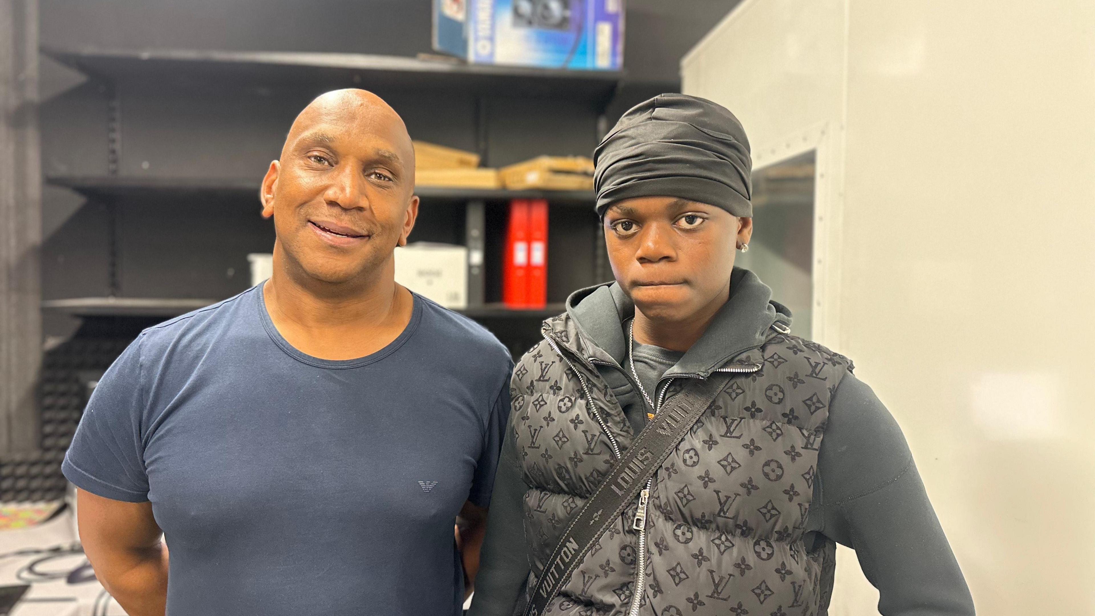 A bald man in a navy blue t-shirt stands next to a young boy wearing a black durag, a dark grey hoodie and a grey gilet with a bag strap over his left shoulder.