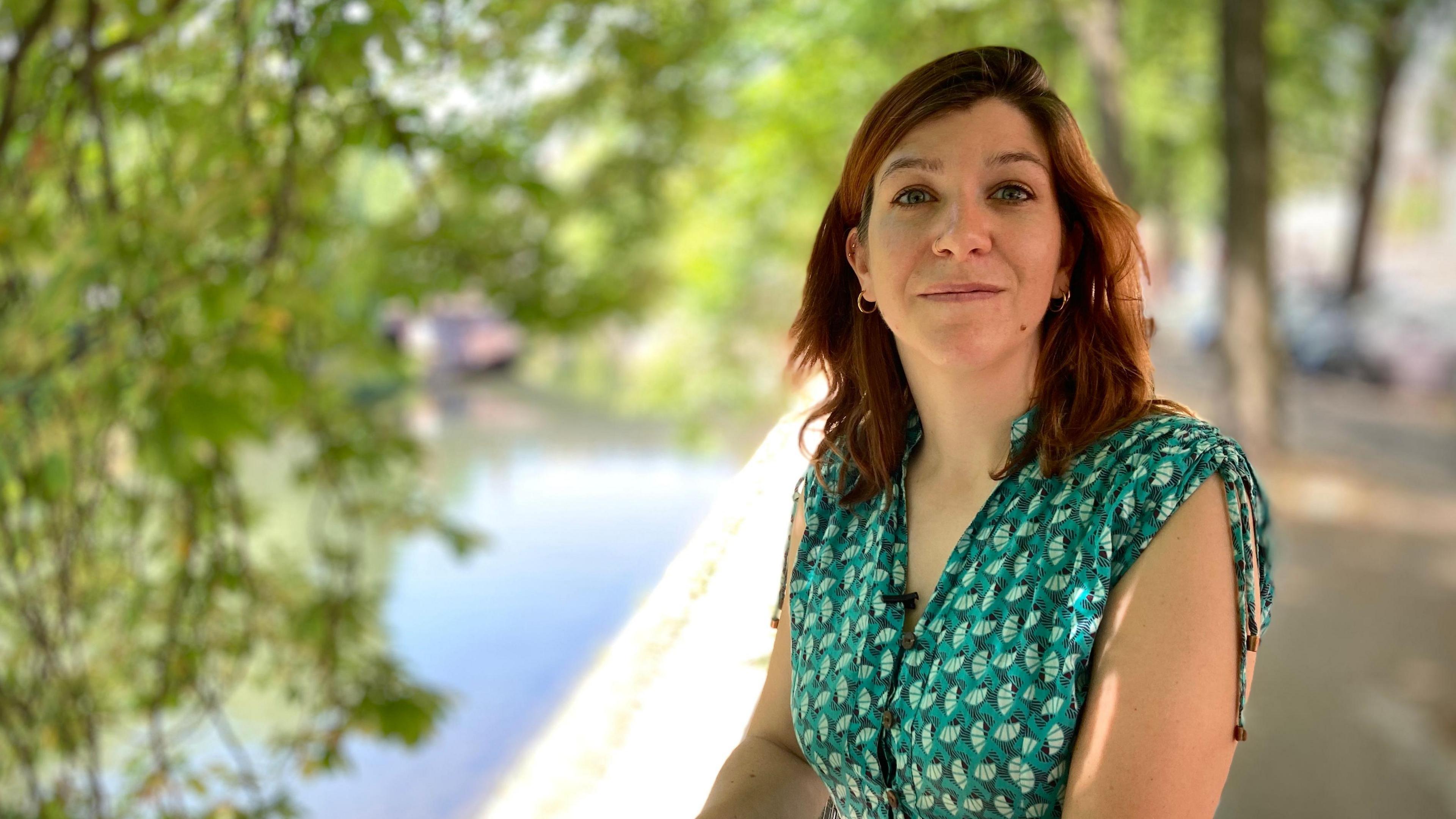 Marie-Hélène Brice is wearing a green patterned, short-sleeved dress. She has straight, shoulder-length brown hair. Behind her are some trees and a stretch of water.