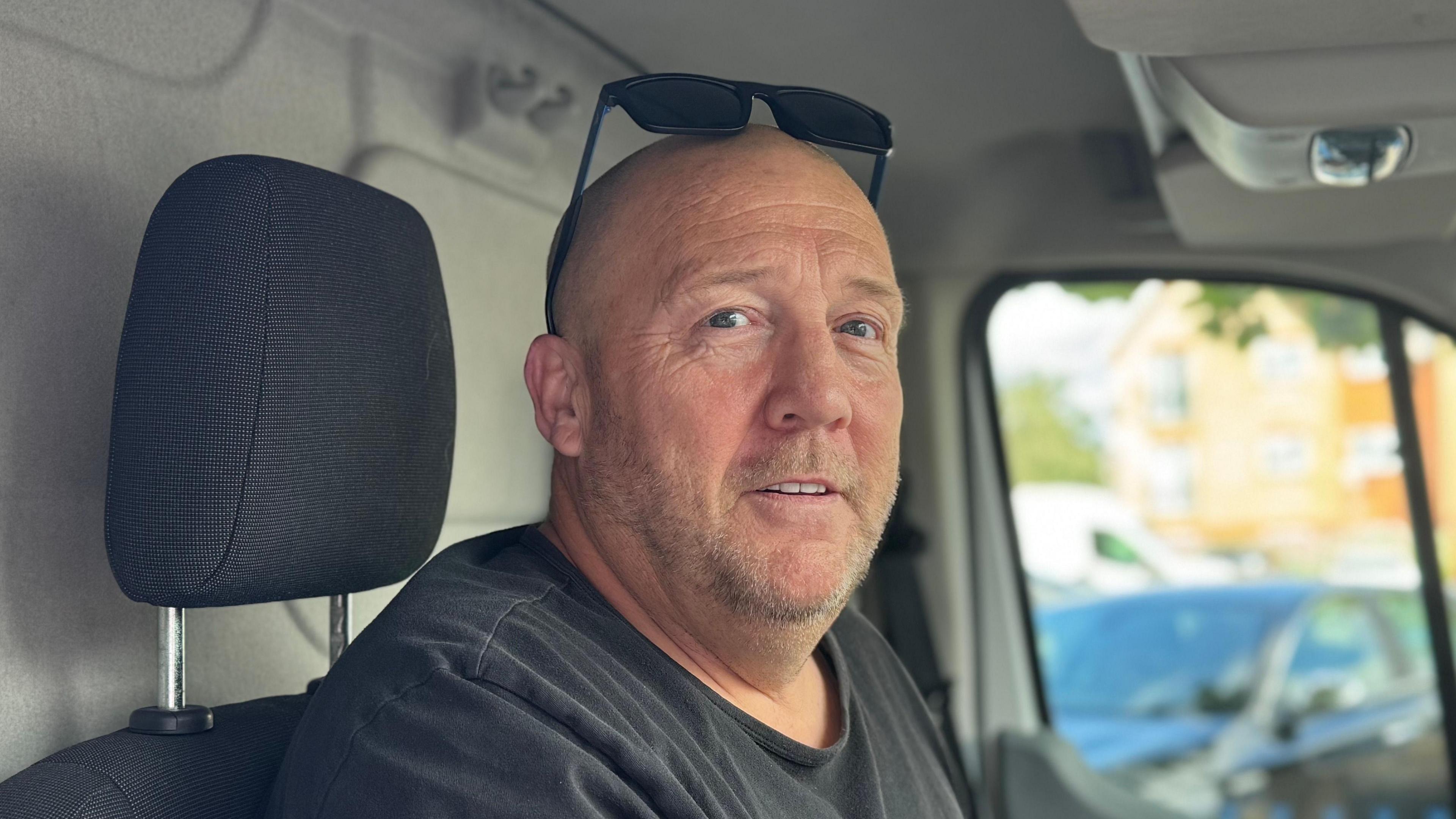 A head and shoulders image of a ma who is wearing a dark T-shirt and sitting in a van. He is looking directly into the camera. His sunglasses are on his head.