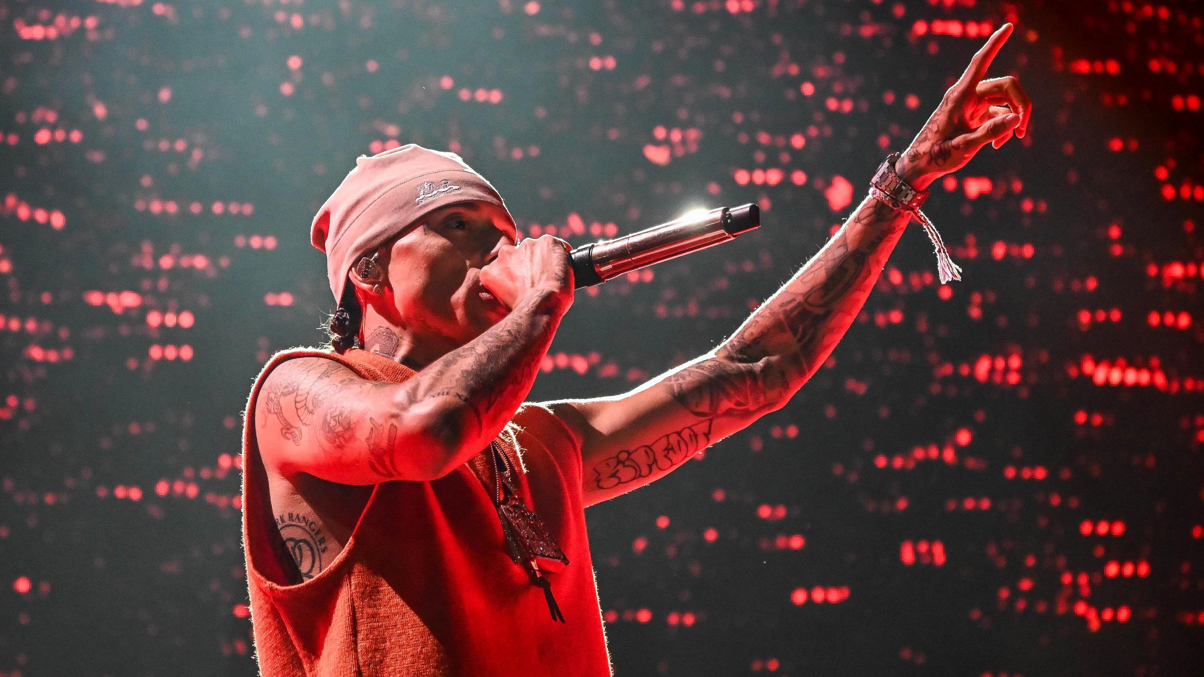 The image shows a person wearing a beanie and a sleeveless top, holding a microphone close to their mouth with one hand and pointing upwards with the other. Their arms are tattooed, and they’re wearing bracelets on one wrist. The background is lit with red lights.