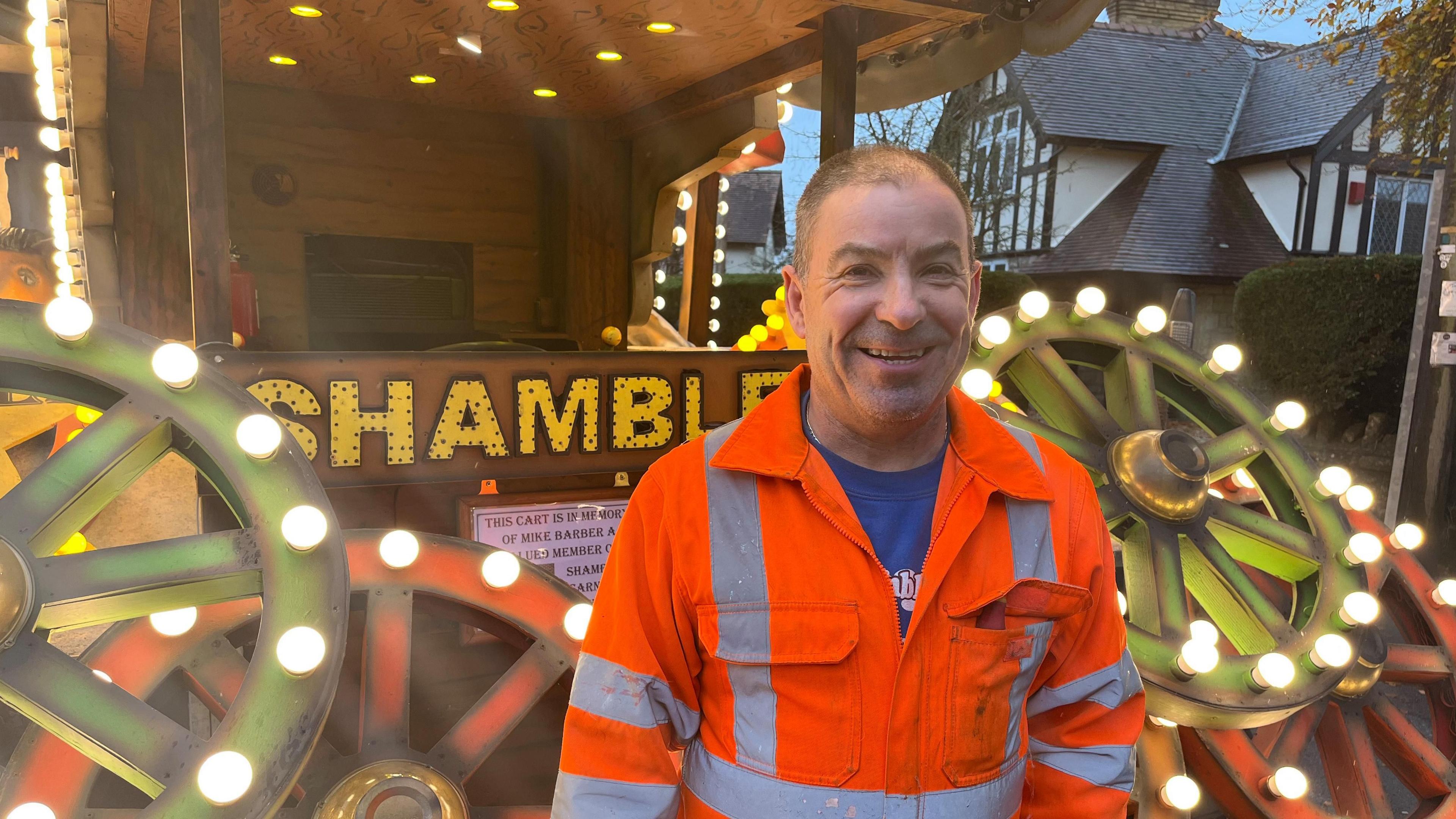 A man in orange hi-vis standing in front of a carnival cart reading 'Shambles' with wooden wheels covered in light bulbs on the front