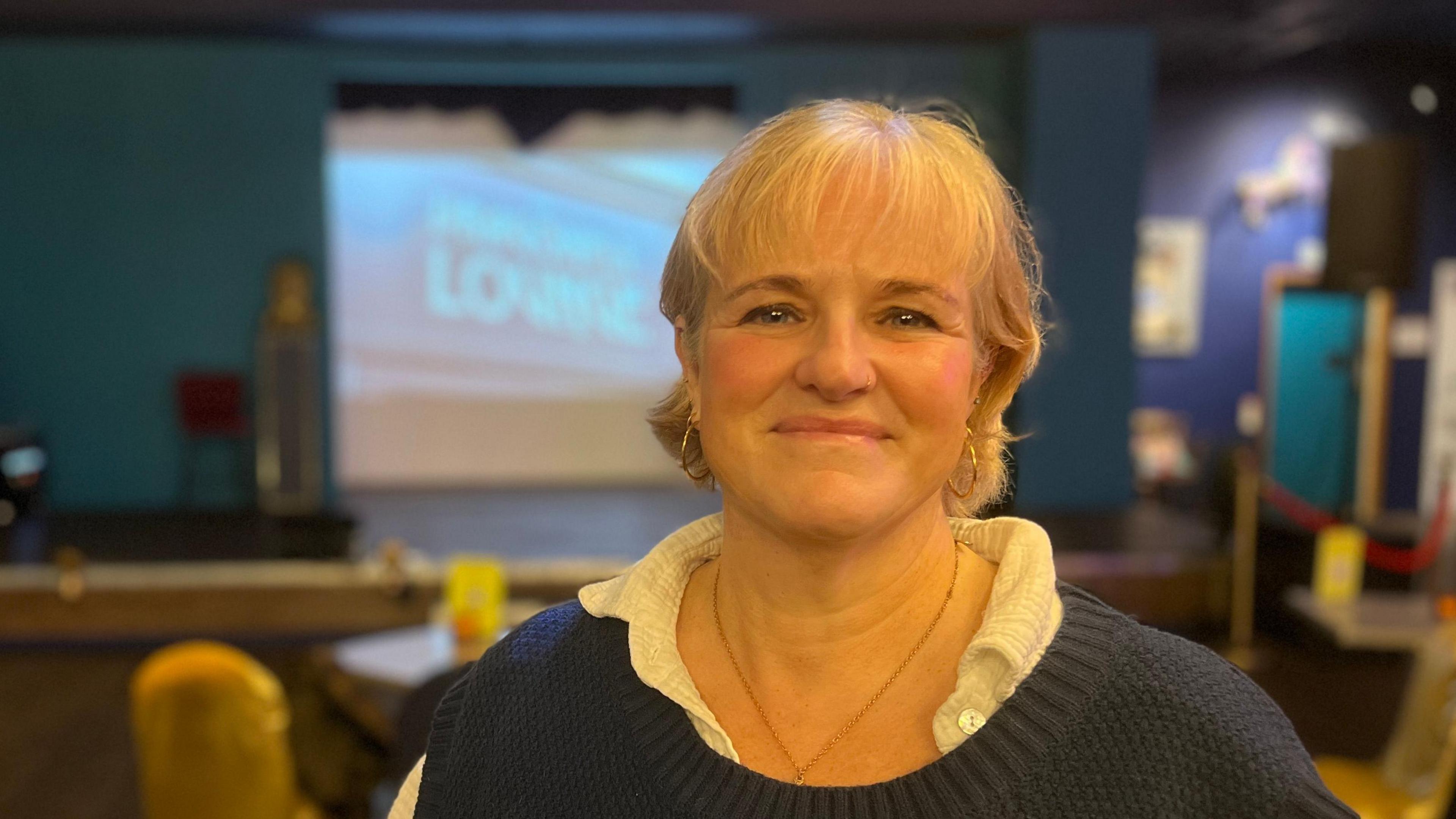 A lady smiling at the camera wearing a white blouse with a blue jumper. She is smiling at the camera. She is in a bar which is out of focus in the background.