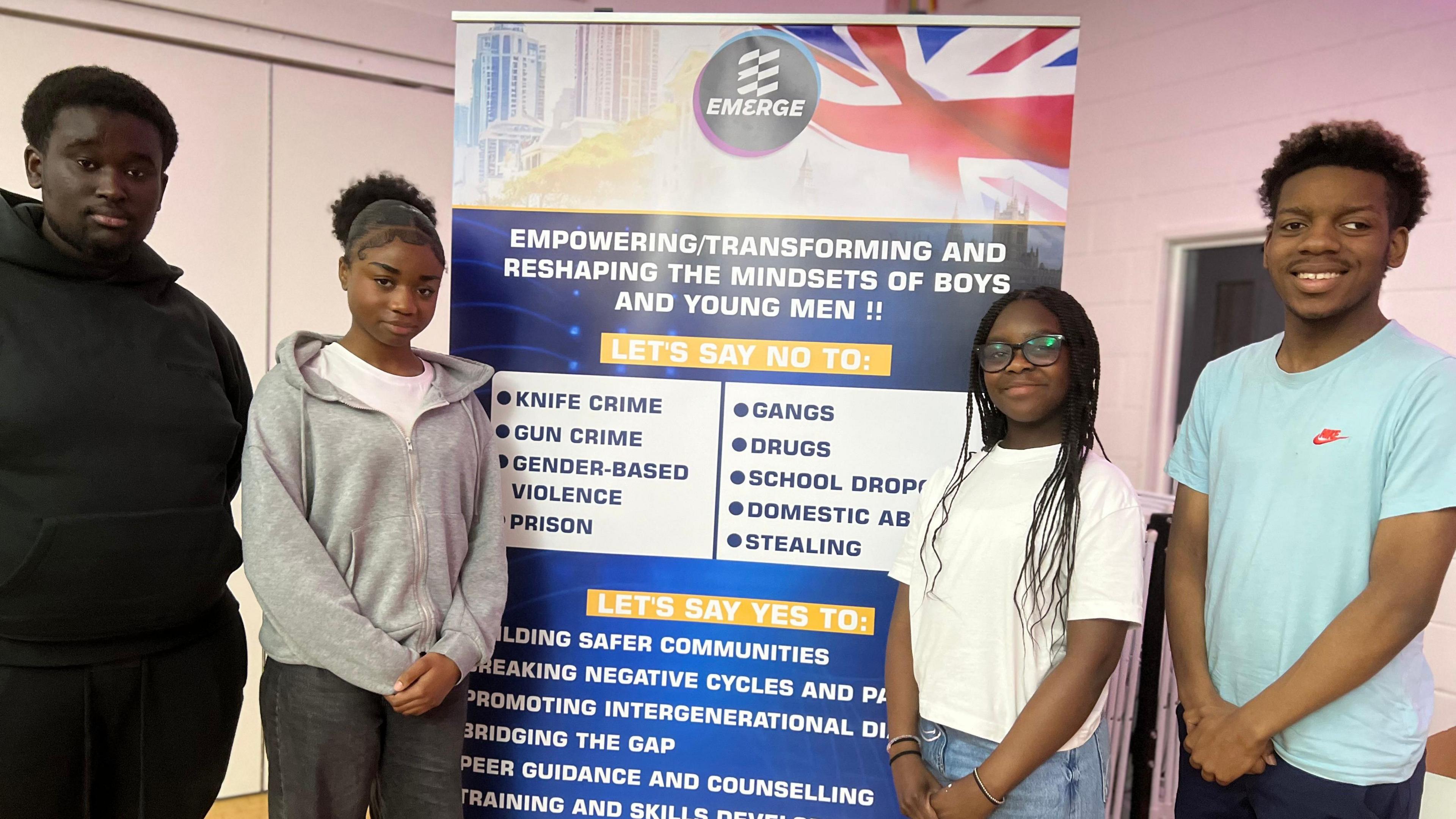 A group of four young people stand in front of a sign promoting the Emerge project, which pledges to "reshape mindsets".