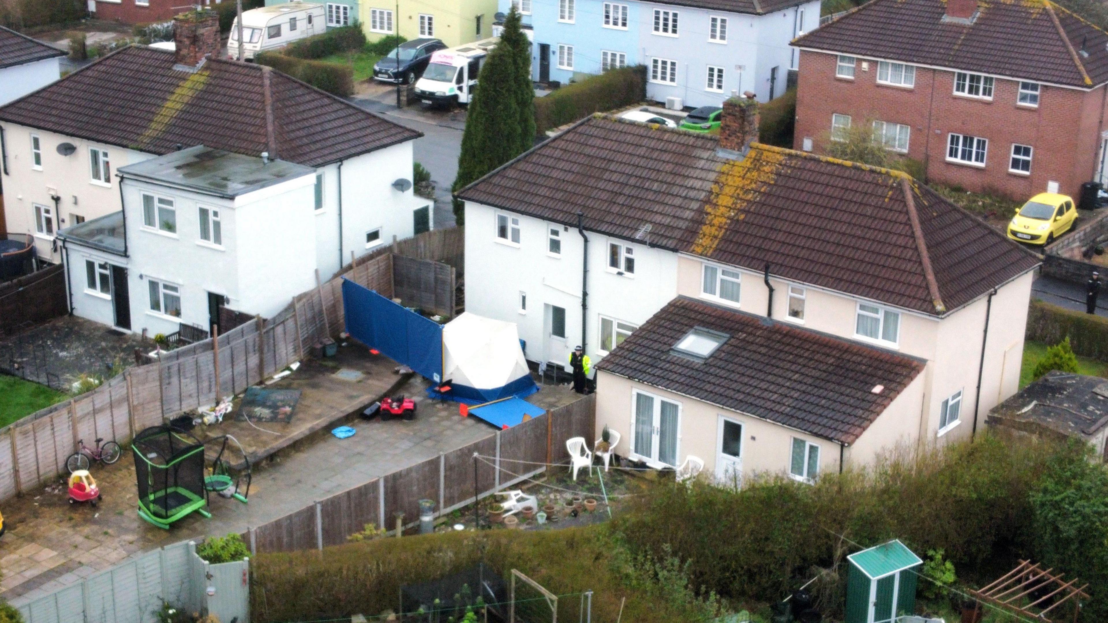 An aerial shot of a house on Blaise Walk in Sea Mills, Bristol. There is a white forensics tent in the back garden. The house is painted white with a brown roof. A police officer wearing a black uniform and a yellow high-vis jacket is standing in the garden. 