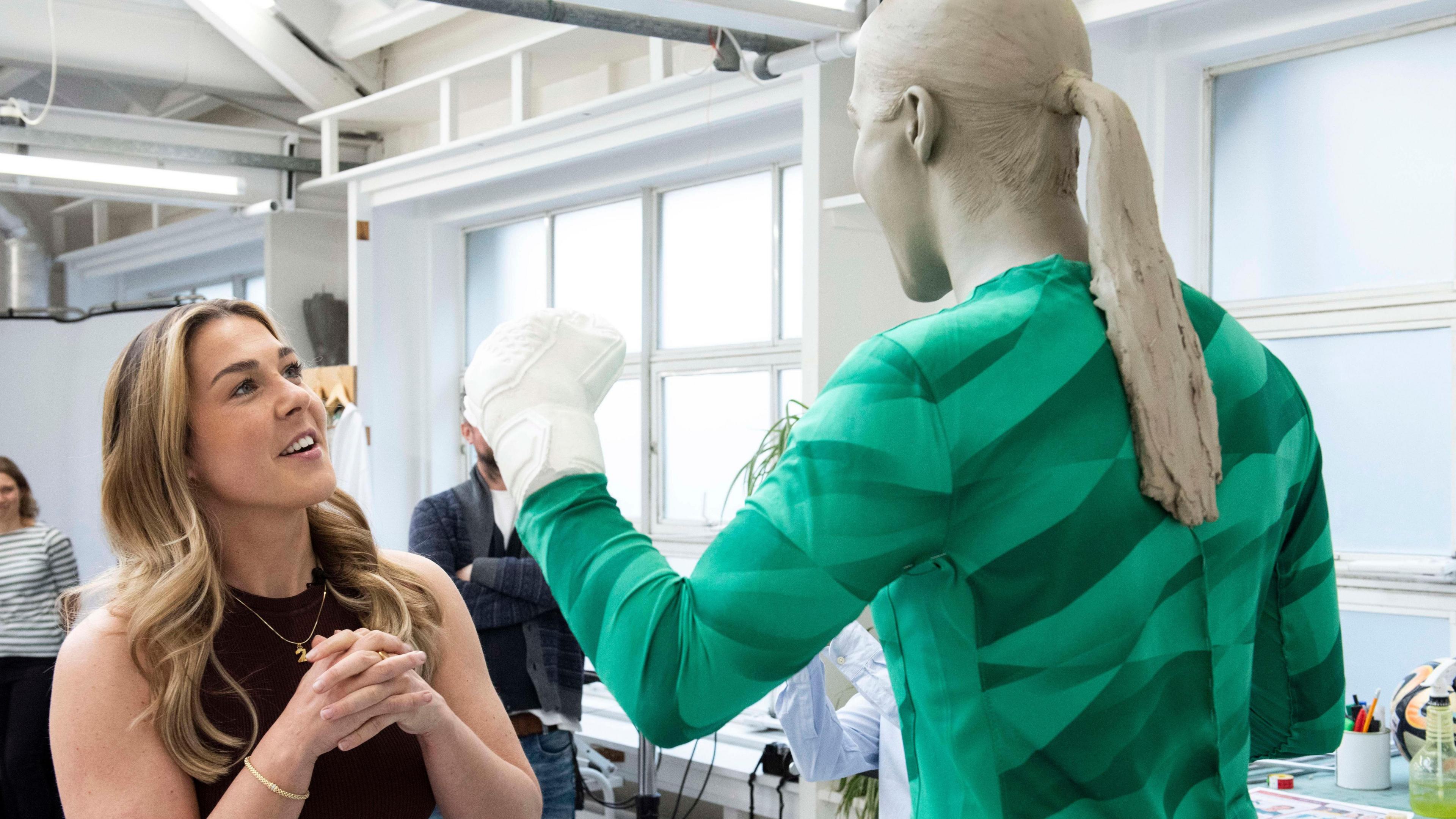 Mary Earps, wearing a black sleeveless dress stands with her hands together as she looks at a waxwork in her likeness wearing a green England goalkeeper jersey.