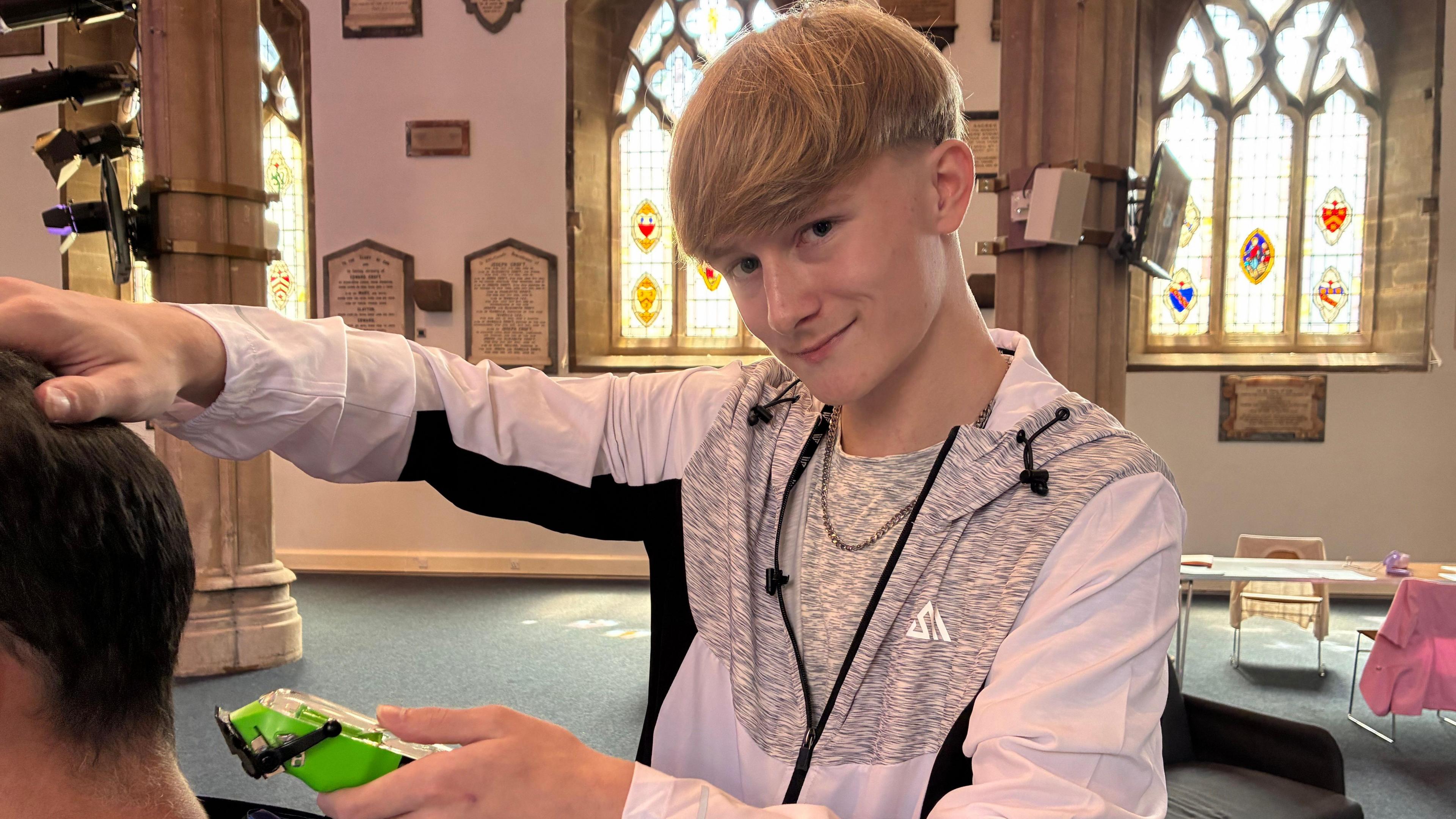 A 16 year old white boy with blonde hair is smiling at the camera while about to cut someone's hair to the left with barber razors. He is in a church in front of three stained glass windows. 
