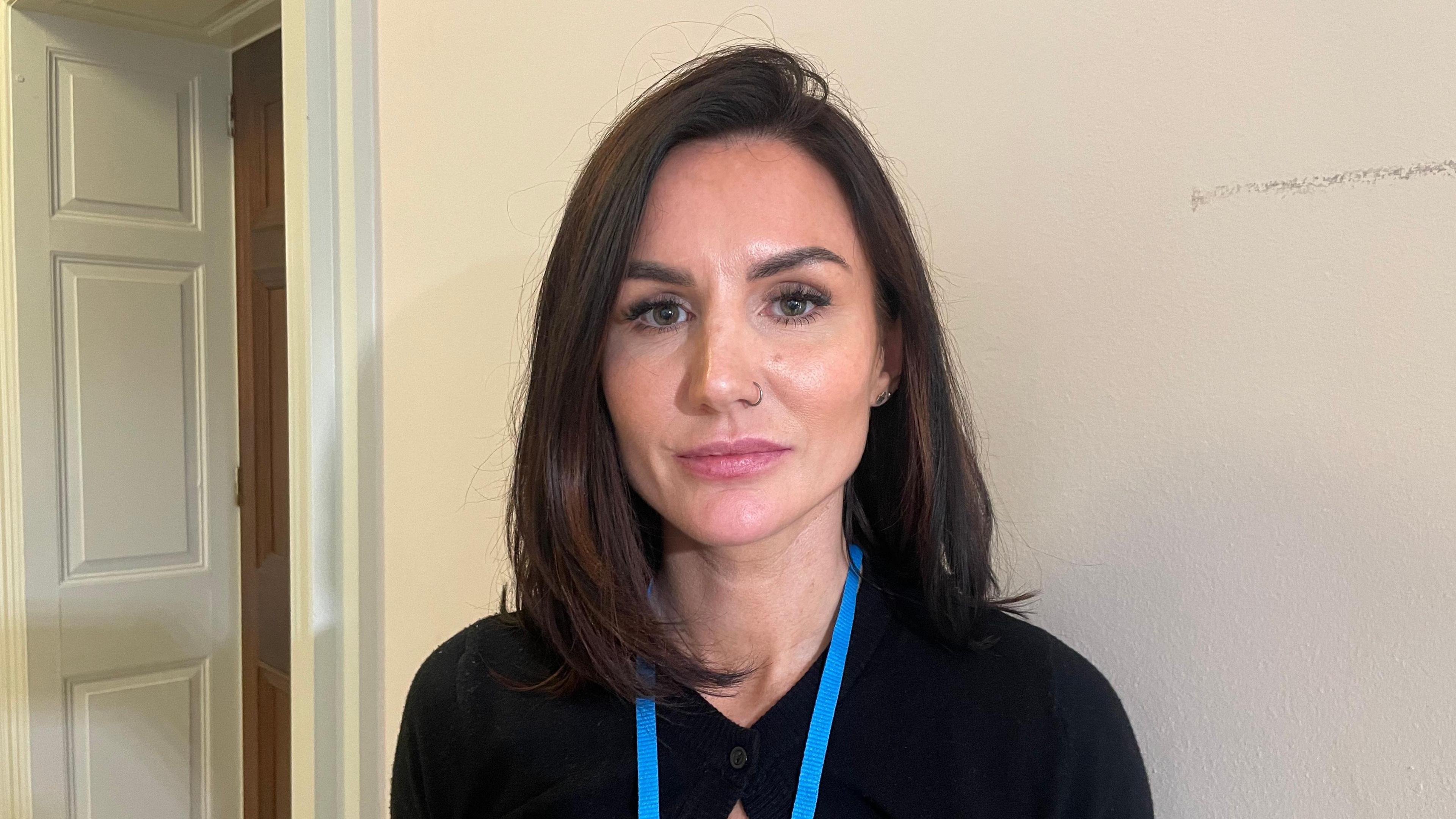 Lisa McCartney is standing looking into the camera in front of a white wall. She is wearing a black top and a blue lanyard