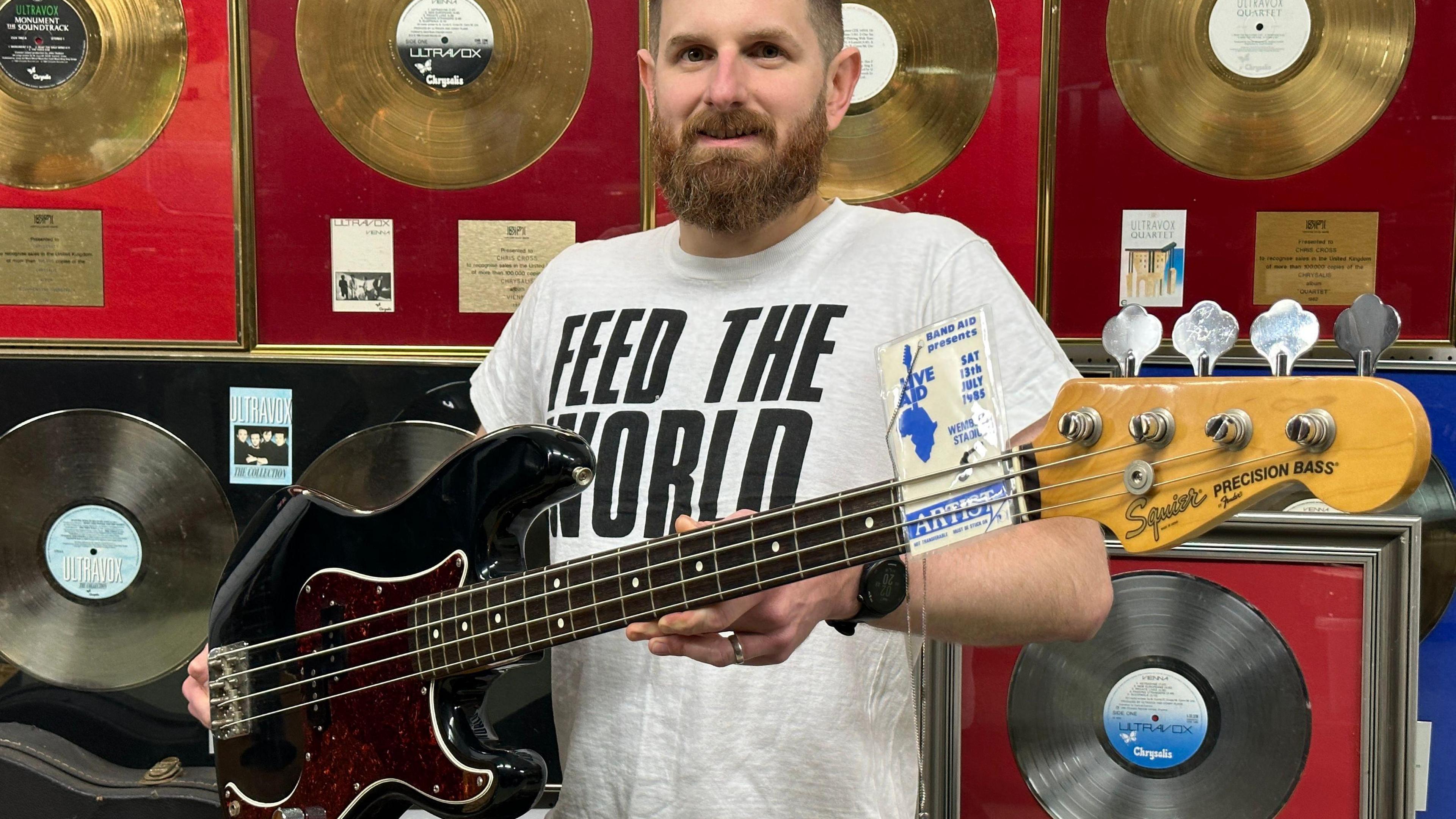 Auctioneer Luke Hobbs from Gardiner Houlgate stands facing the camera holding a black bass guitar which was played by Chris Cross at Live Aid. Mr Hobbs has a beard and is wearing a white T-shirt with "Feed the World" in black letters on it