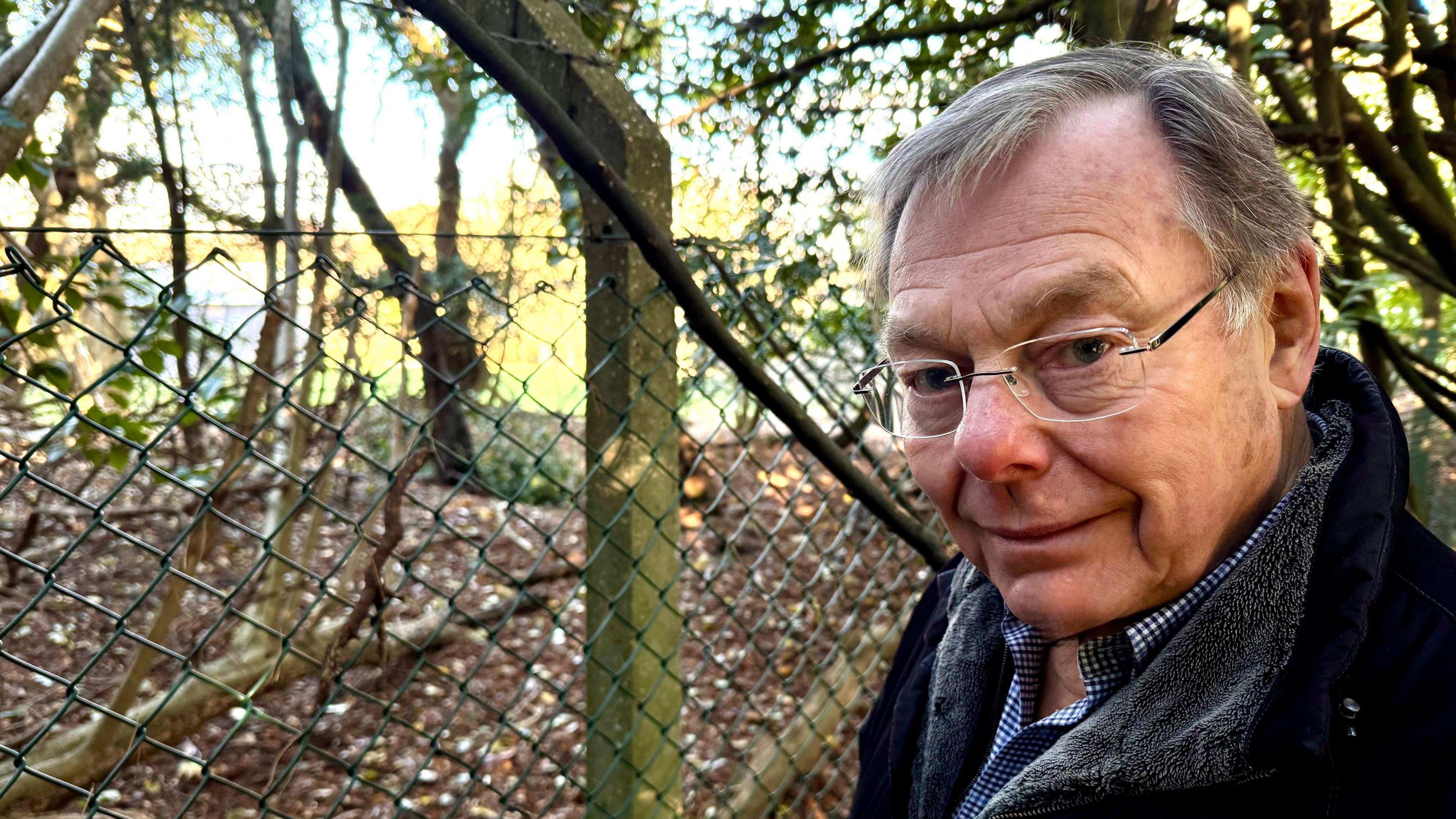 A man in glasses and a dark coat stands in front of a barbed wire fence. Behind the fence a building belonging to the army camp can be seen.