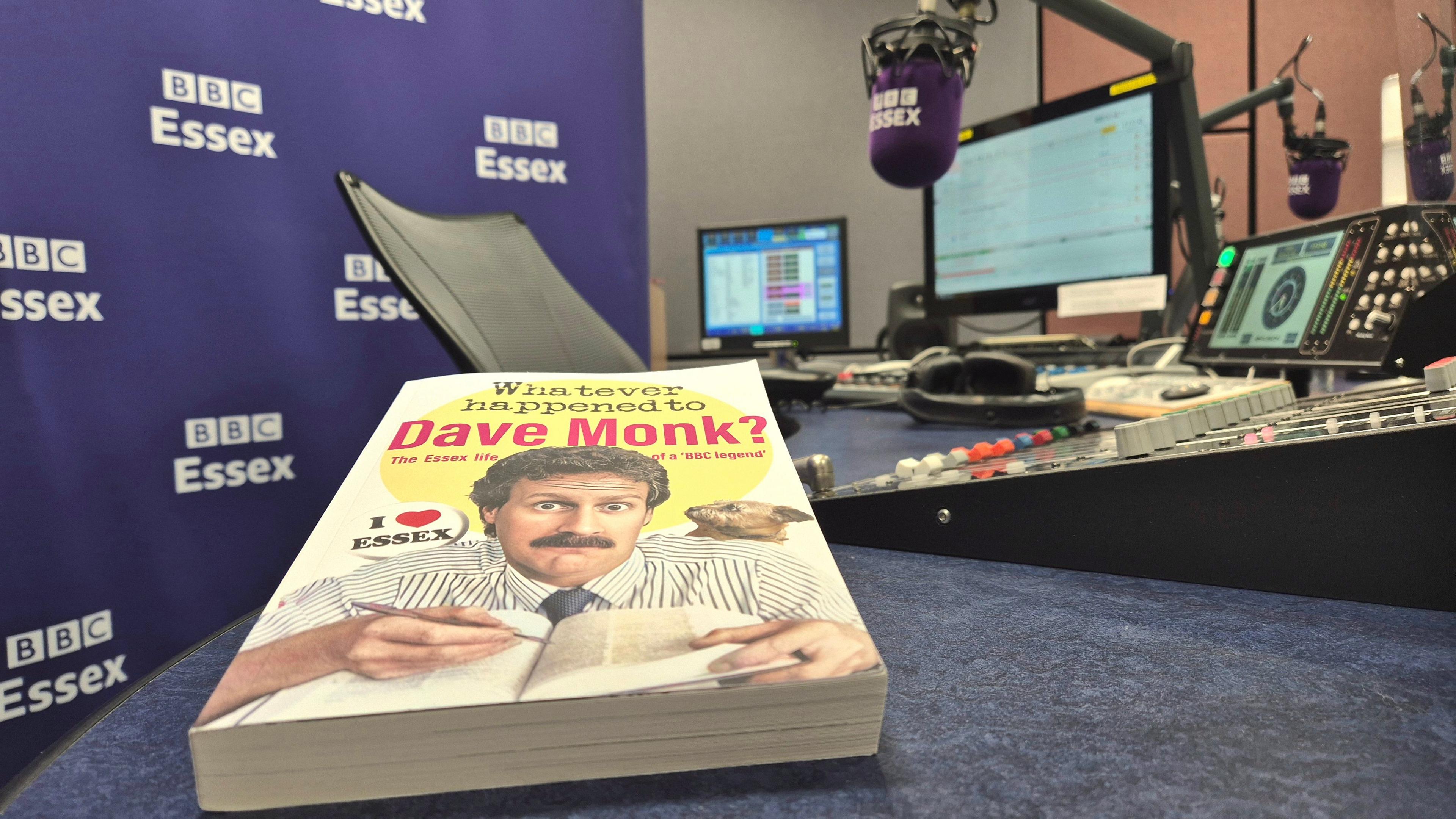A book entitled "Whatever happened to Dave Monk?" sits on a desk in a radio studio. There are microphones, headphones and mixing desks in the background, as well as a large purple background with several white BBC Essex logos on it.