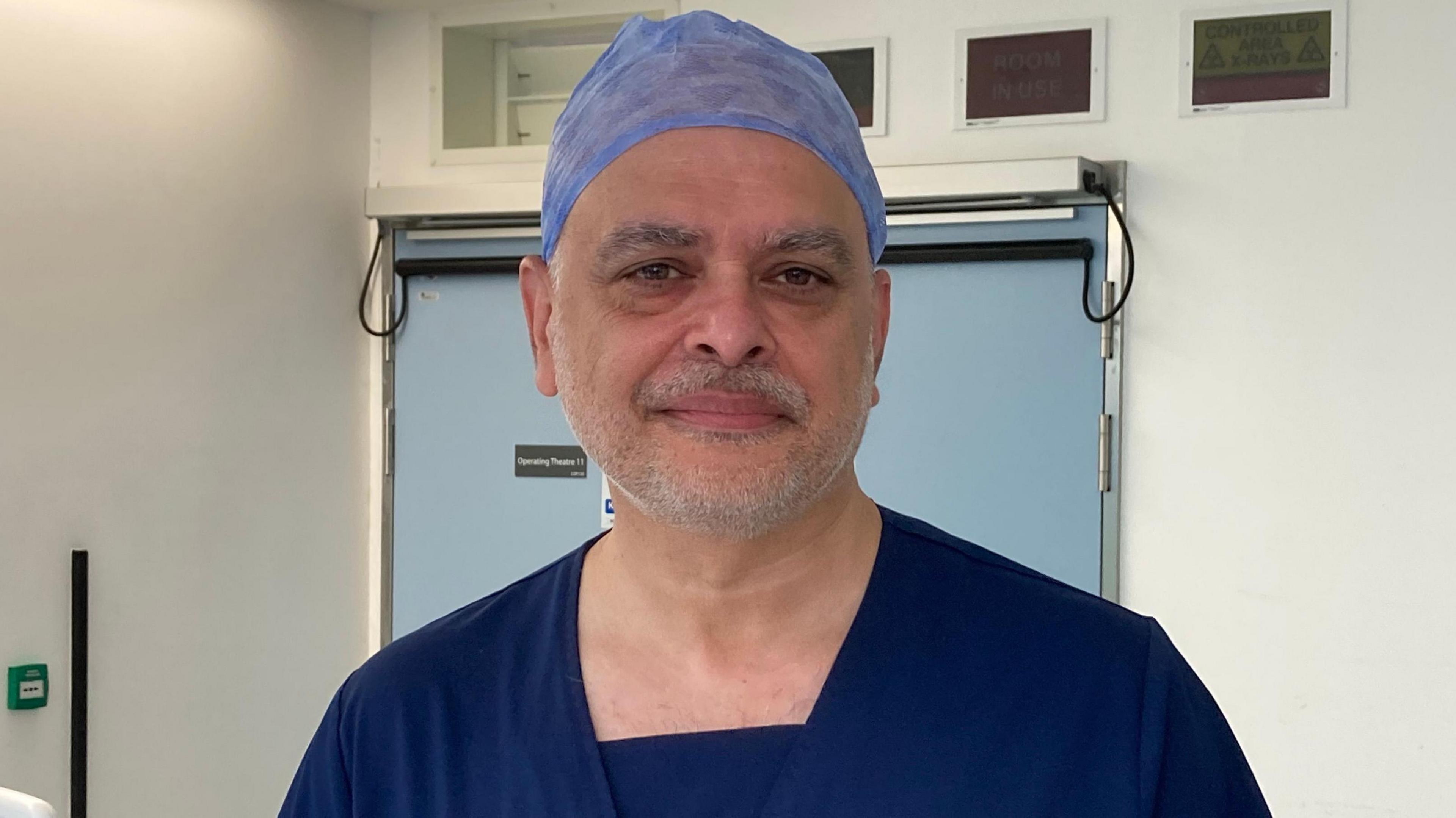 A surgeon is looking into the camera. He's wearing navy scrubs and a light blue covering on his head.