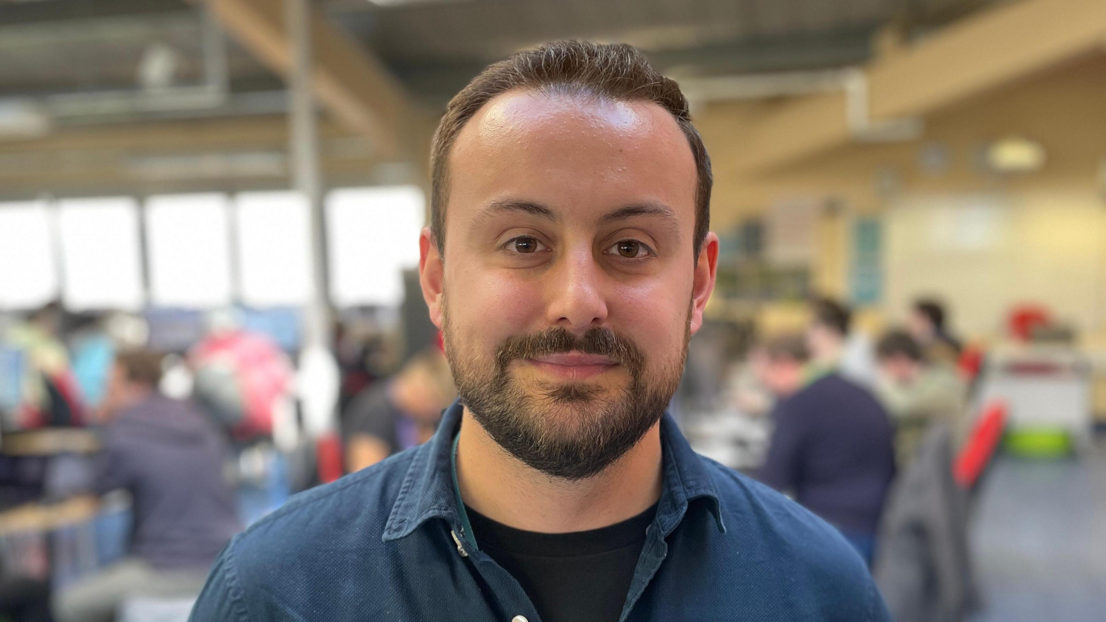 Course leader Michael Farrow-Killick is wearing a navy blue shirt and black T-shirt. He is standing in the middle of the games development classroom at Cambridge Regional College. Students are out of focus in the background but you can tell they are looking at computer screens.