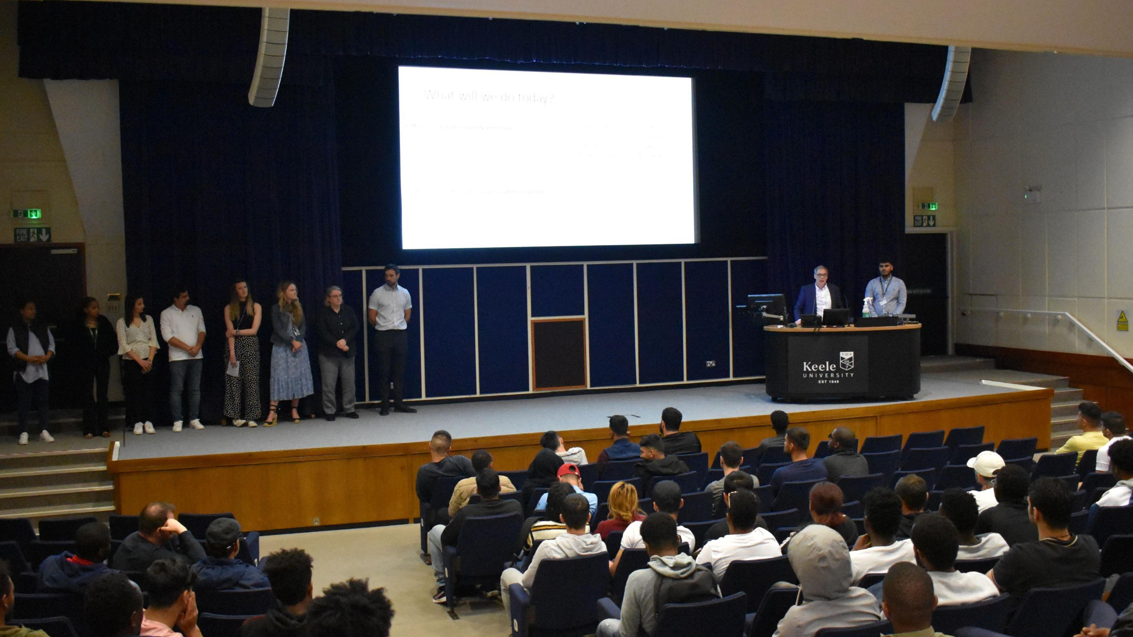 A lecture hall full of people sit in front of an illuminated screen on a wall. two people stand talking behind a plinth that reads "Keele university". to the left, eight people stand on stage, waiting to speak