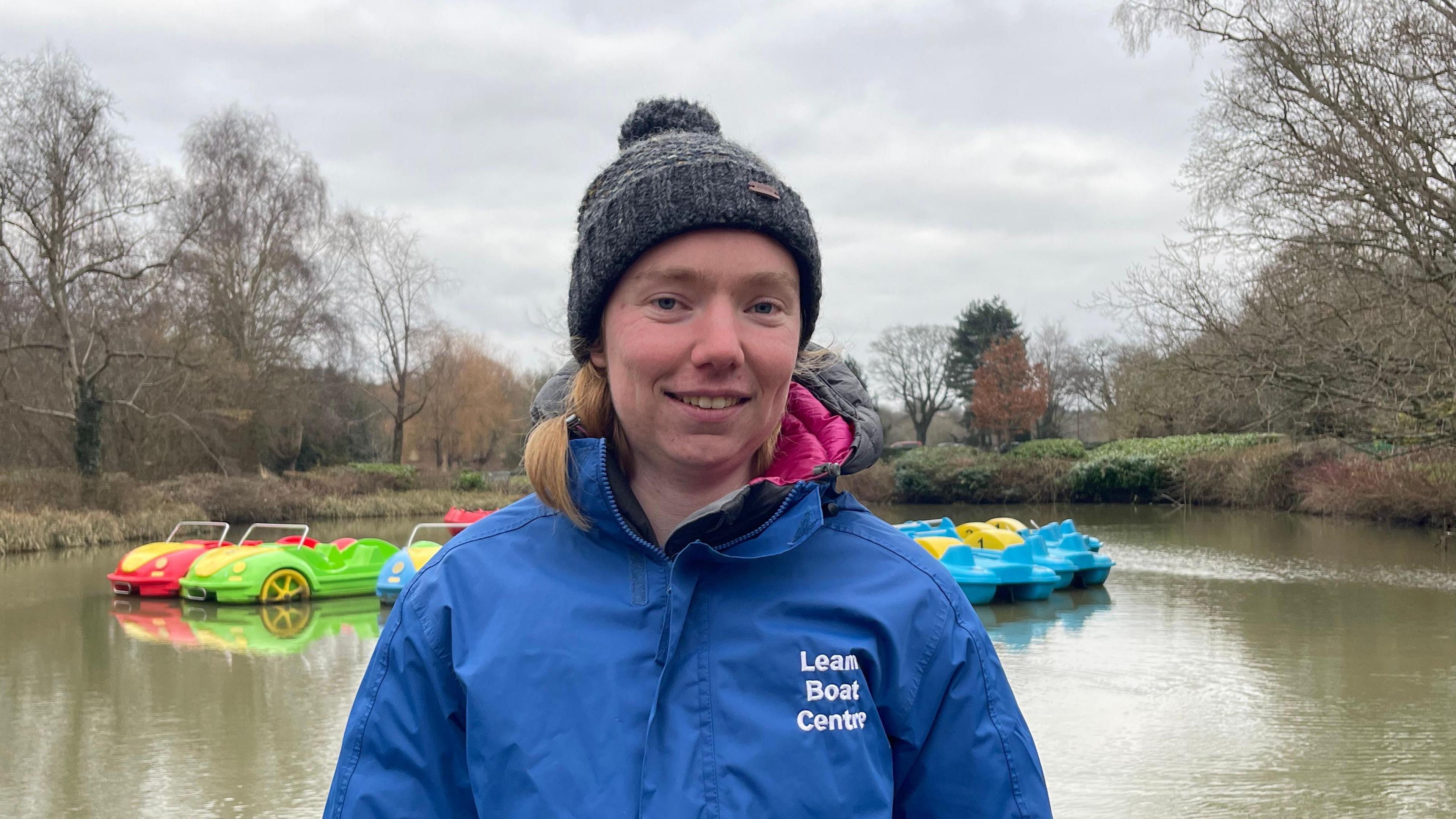 Sophie Dollar in a blue coat stood in front of a body of water with pedalos shaped as cars in the background