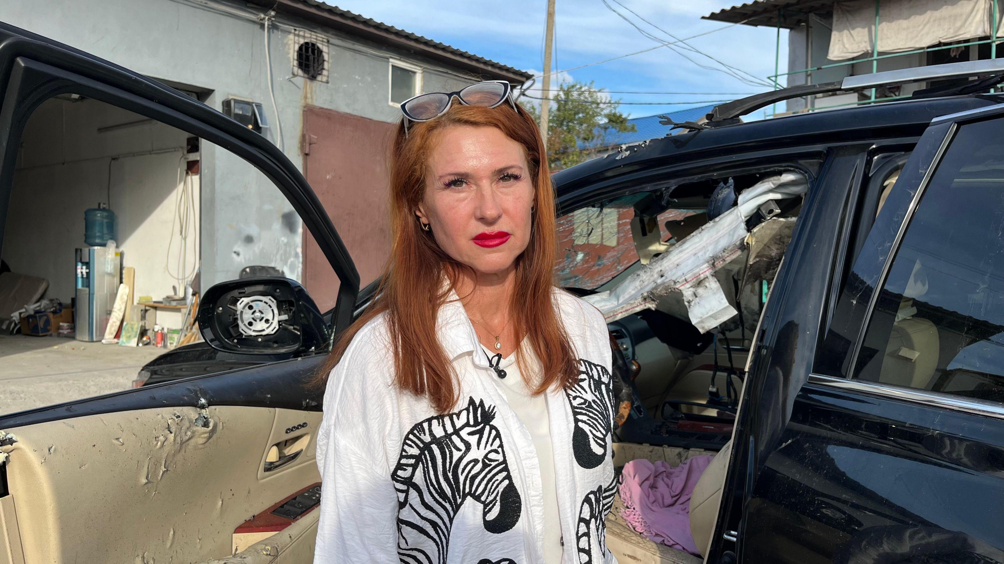 Olena Kryvchun, a woman with red hair and her glasses perched on her head, wearing a white shirt with zebras on it and bright red lipstick, stands by her car. A smashed windscreen, collapsed roof and an interior torn by shrapnel are visible.