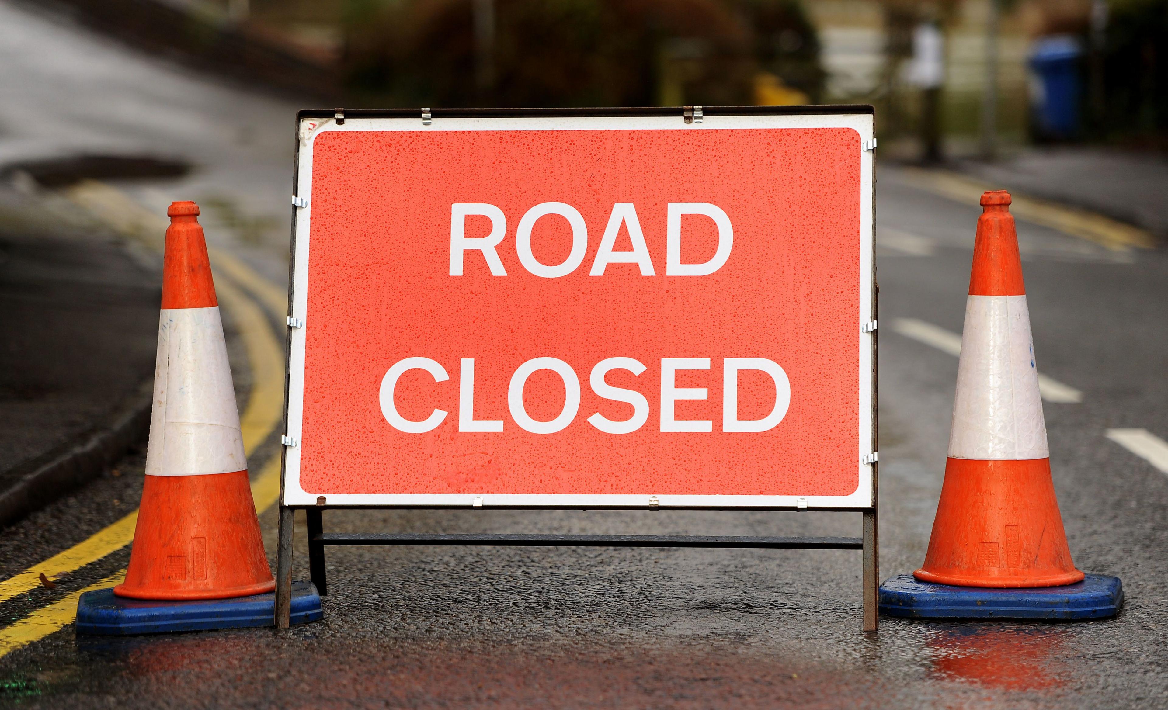 A red 'road closed' sign with white writing is pictured on a road with two traffic cones on either side. Behind and beside it, you can see white and yellow road markings.