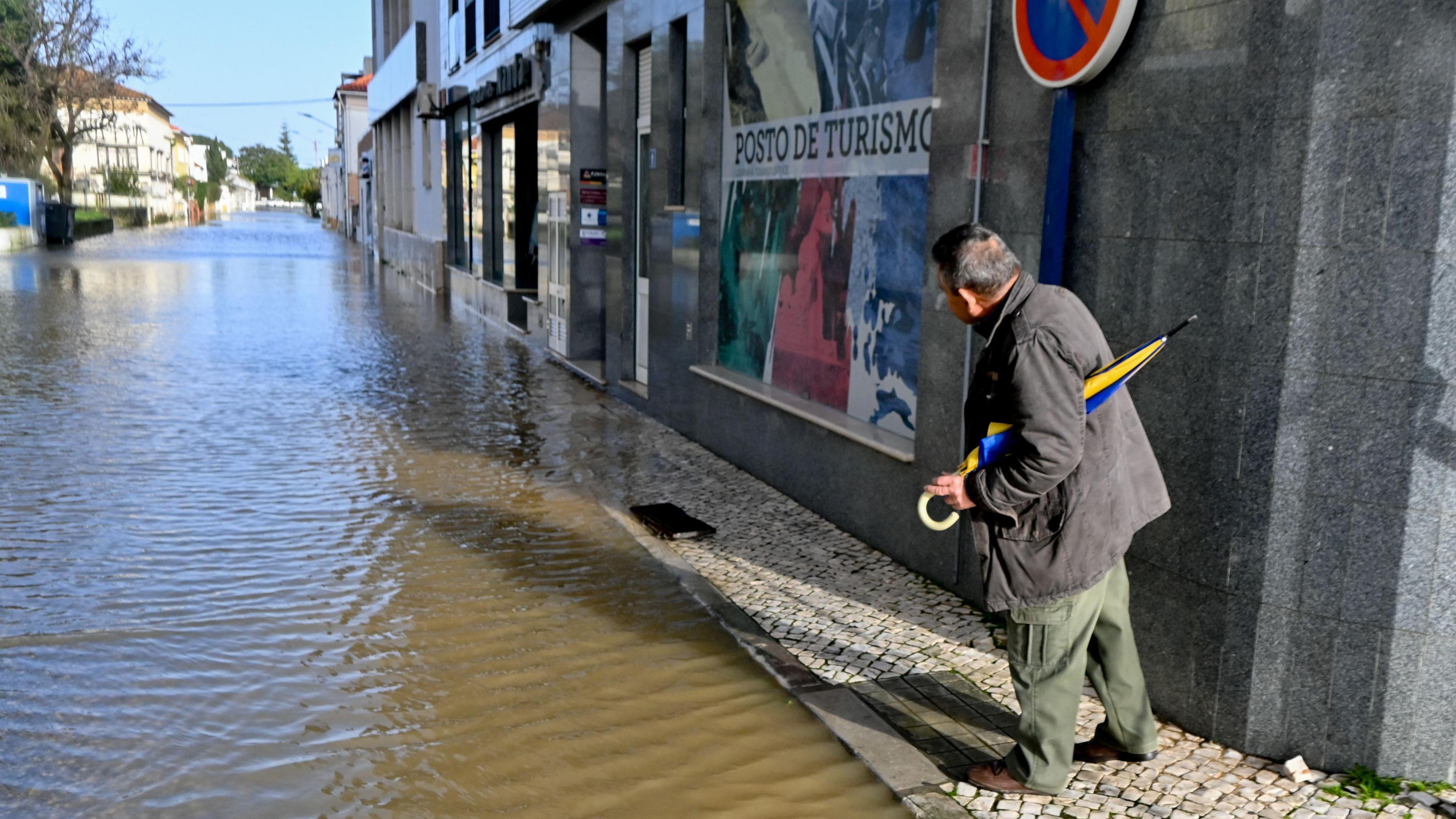 A man holding an umbrella stands on a pavement looking down a flooded street.