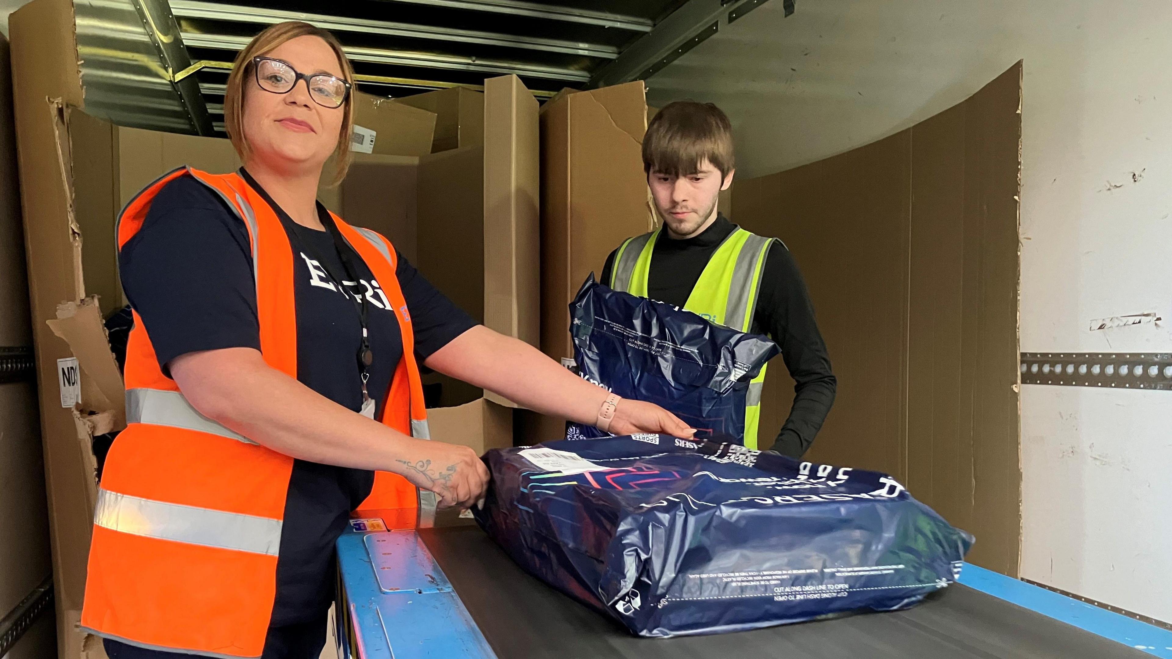 Lauren Smith, wearing an orange hi-vis waistcoat, is helping remove parcels from the back of a lorry and placing them on a conveyor belt to be sorted. A colleague in a yellow hi-vis waistcoat works alongside her