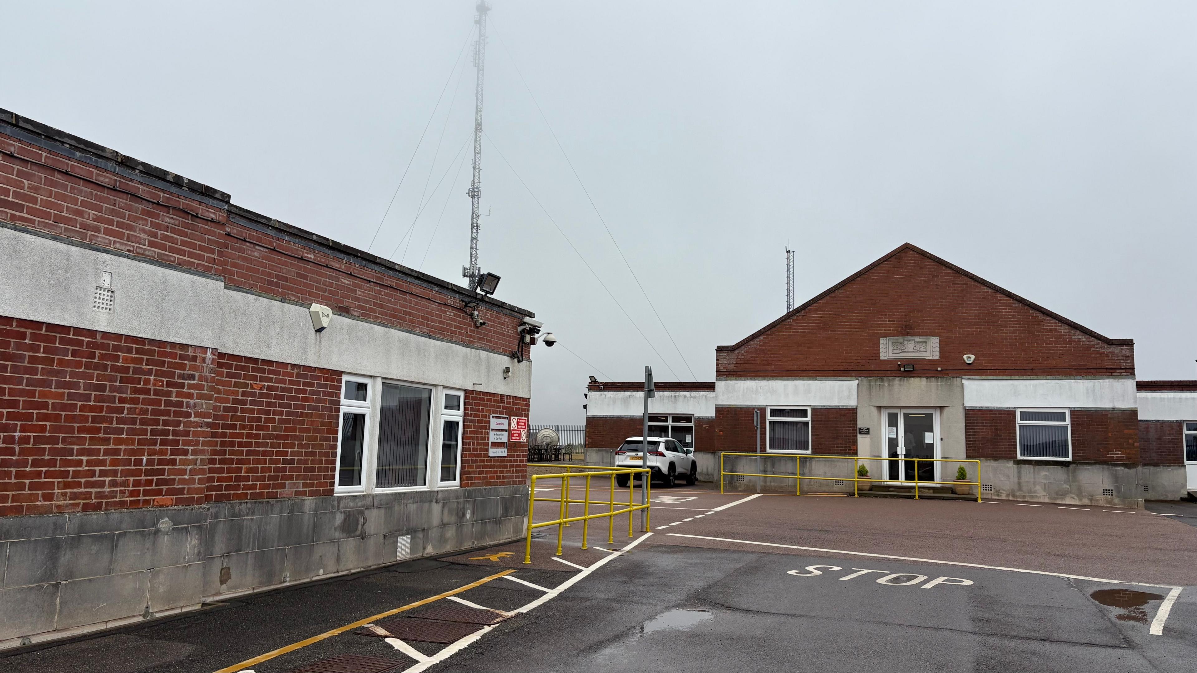 Two single-storey brick buildings. One has a flat room and the other has a pitched roof. There are white-framed windows in both buildings and a door is visible in the furthest building. There is a white square painted on the ground with the word STOP in white. A car is parked in the distance. There is a metal radio mast behind the building on the left.