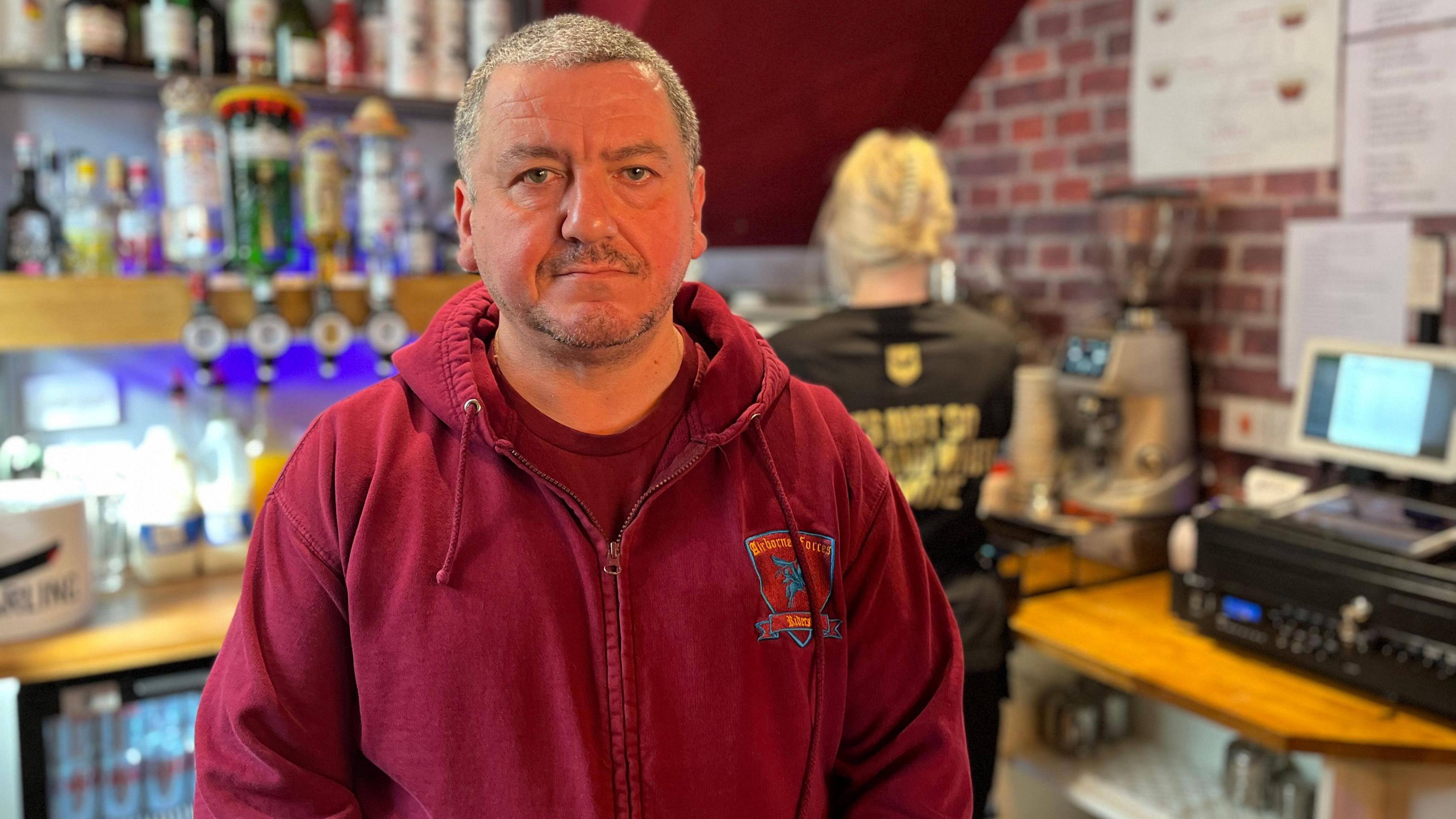 Andrew Hrrison standing in front of a bar with a till to the right and a member of staff in the background