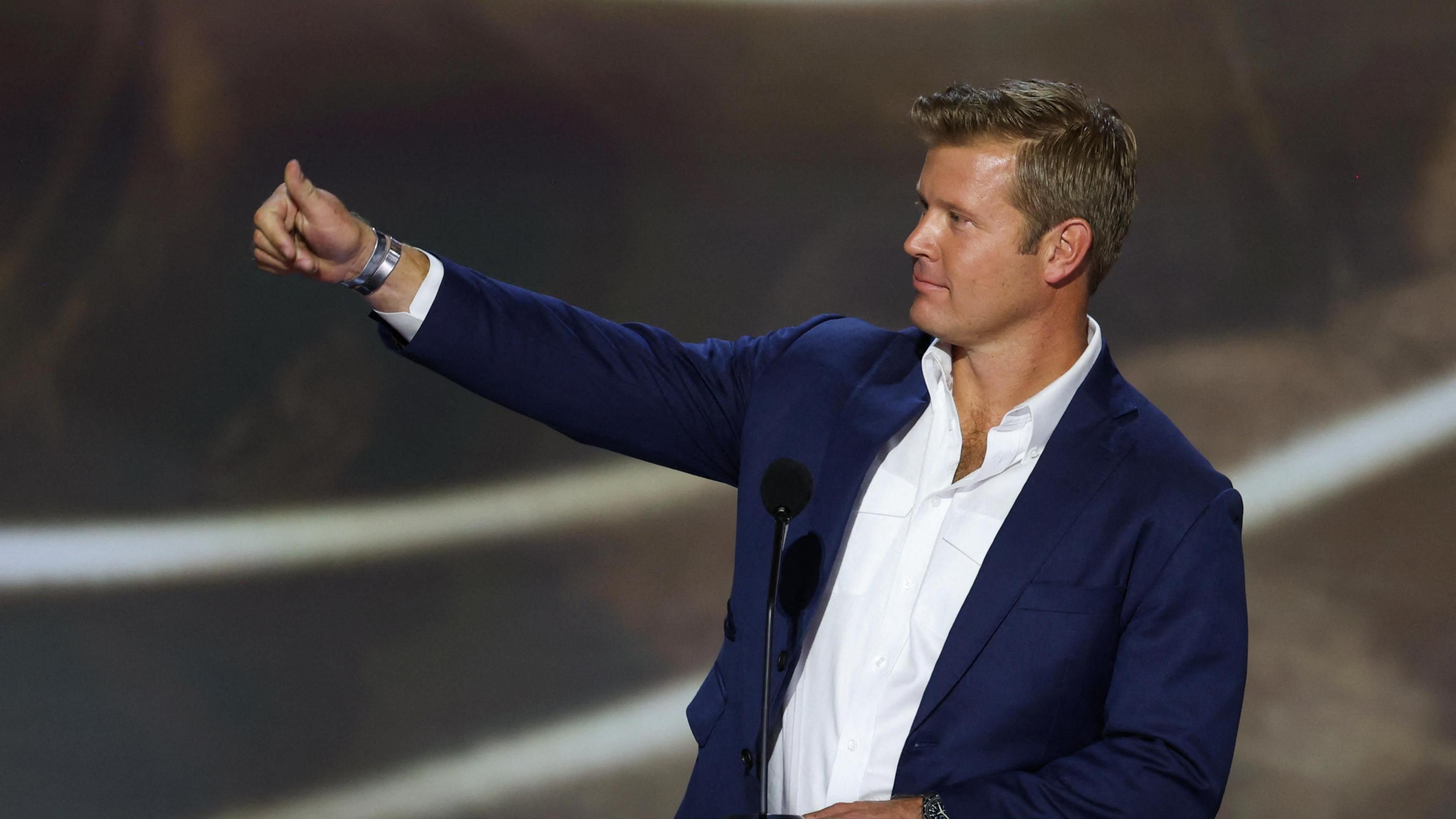 Tim Sheehy gives a thumbs up as he looks to the left wearing a blue jacket and open-necked shirt, at the Republican National Convention in July