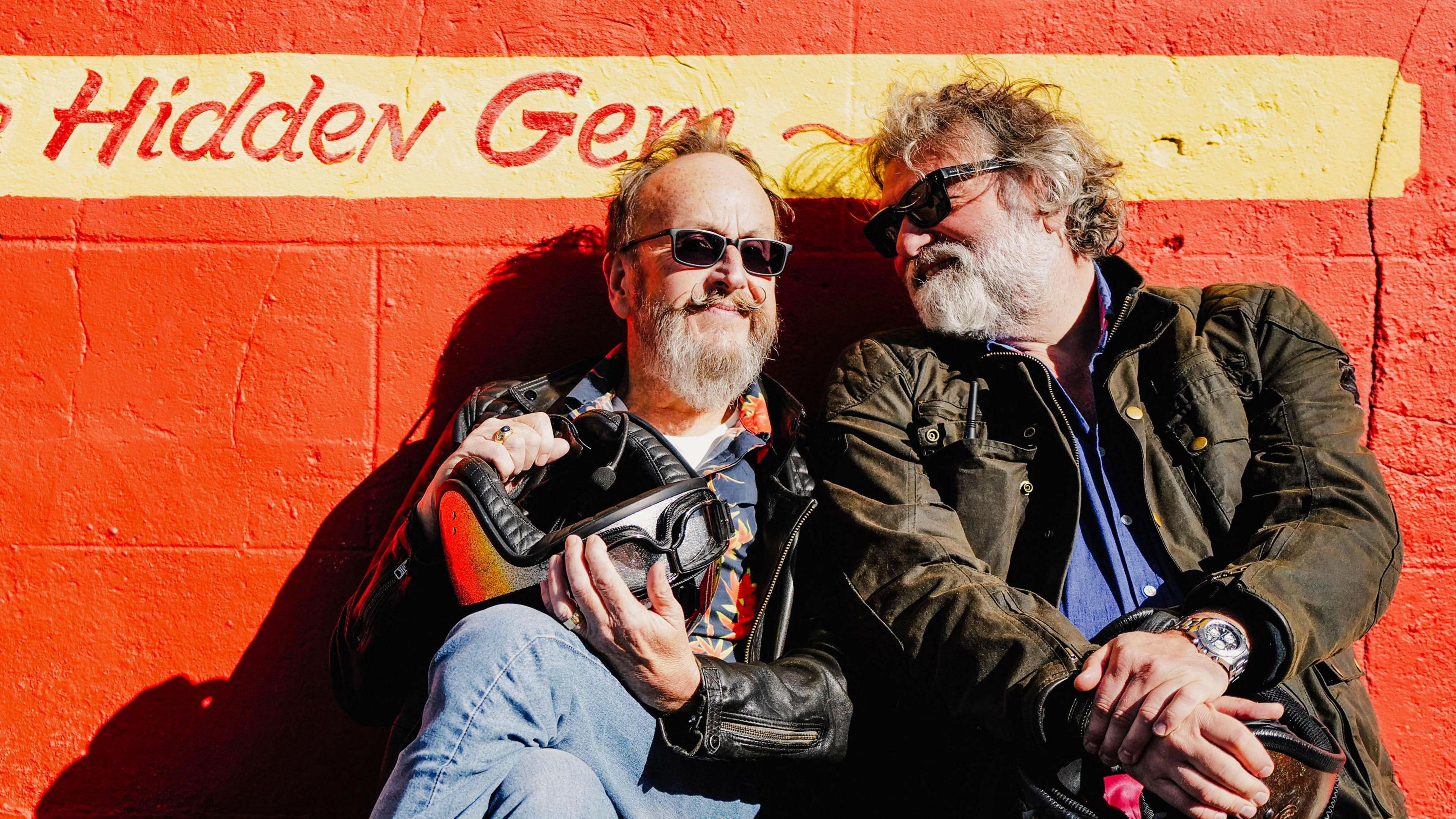 Dave Myers and Si King sit in front of a painted orange wall. They are both wearing biker jackets and have sunglasses on. Dave is smiling contentedly and Si is looking at Dave with fondness. Behind them, written on the wall, is a sign that reads "hidden gem".