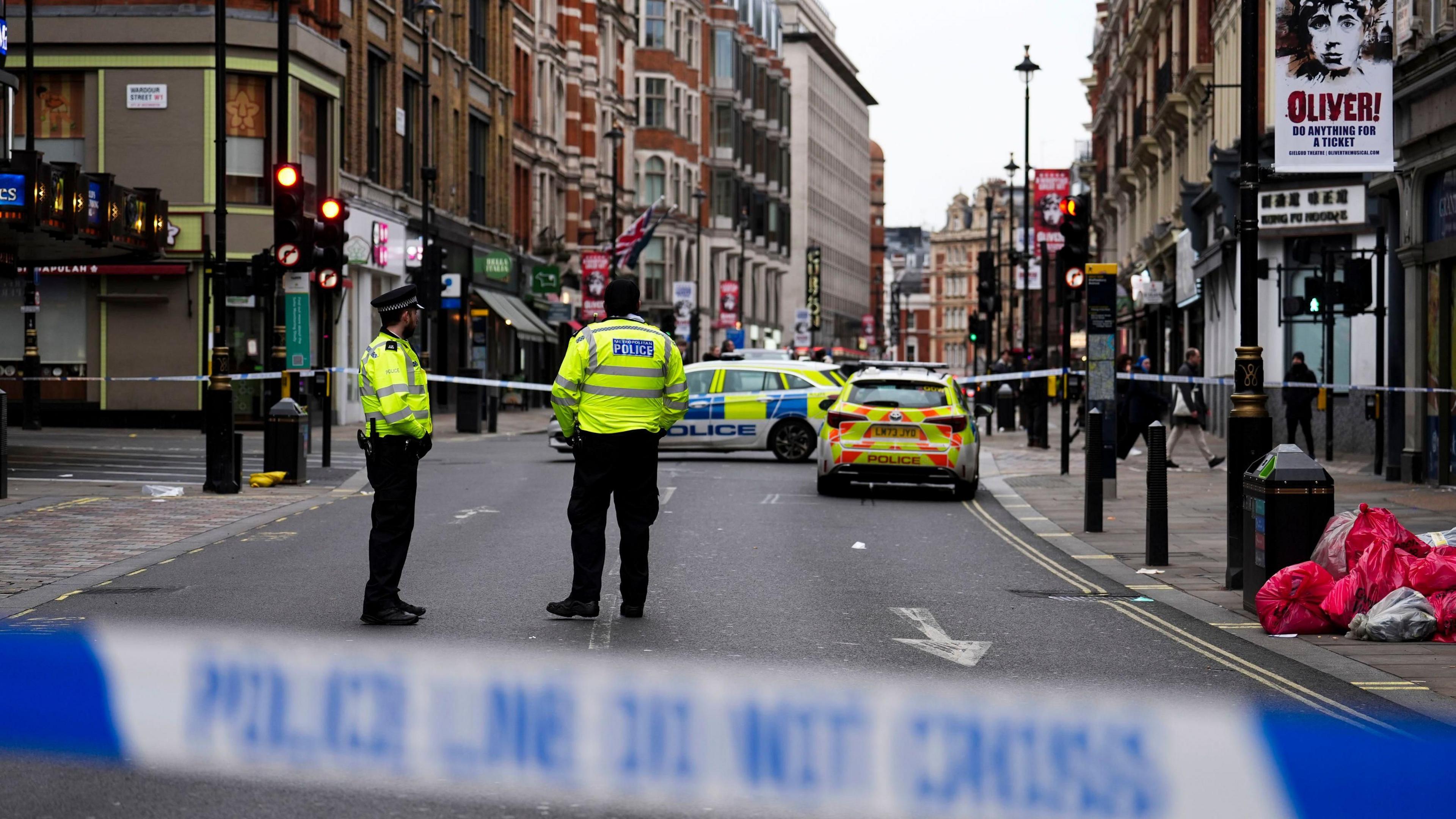 The scene on Shaftesbury Avenue, showing a close-up of blue and while police tape, with two uniformed police officers standing in the street in front of another line of tape and two police cars