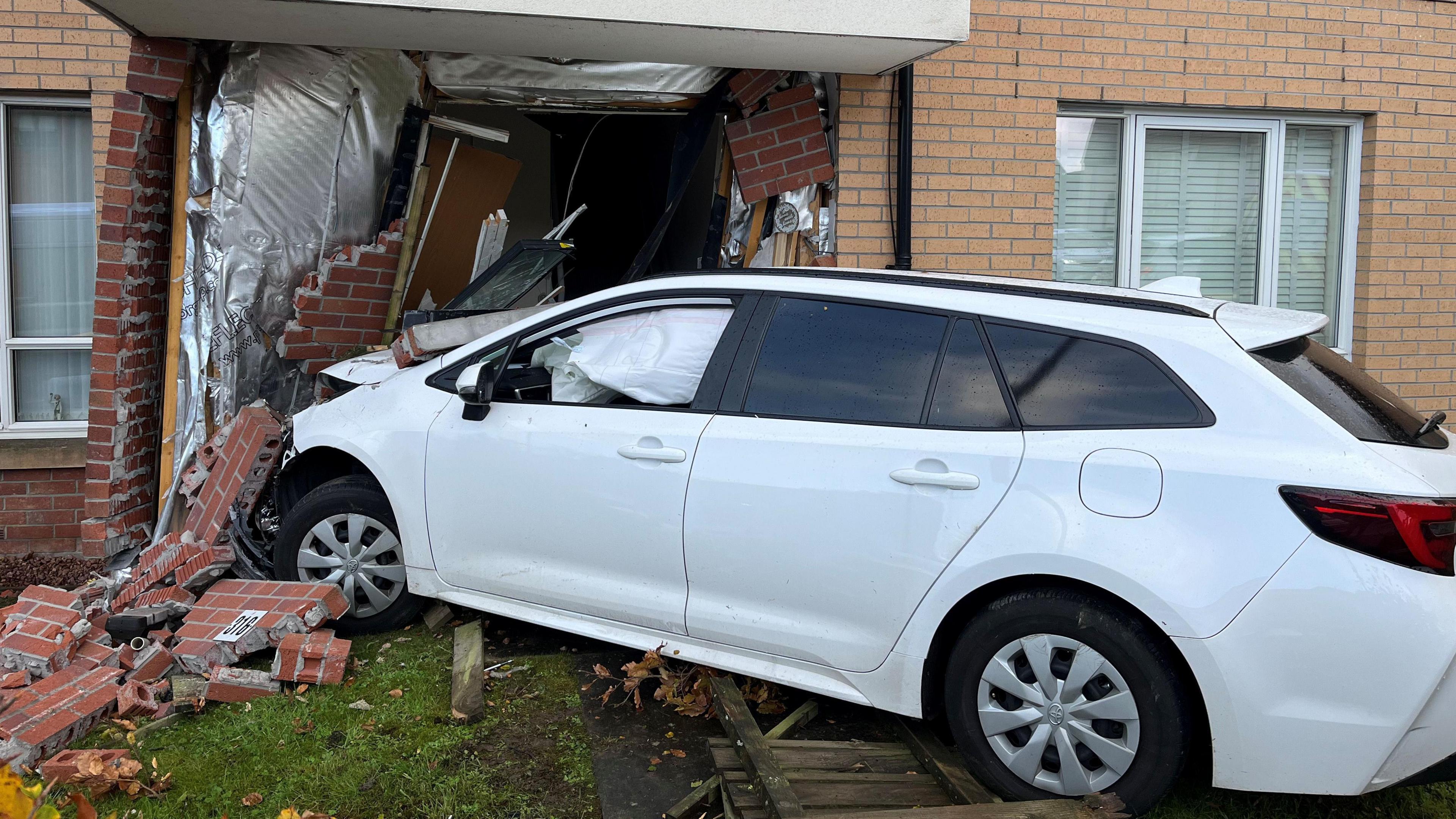A white car that has crashed into the front of a house. There are bricks lying around the car and the front garden , and the car's inflated airbags can be seen through the windows.