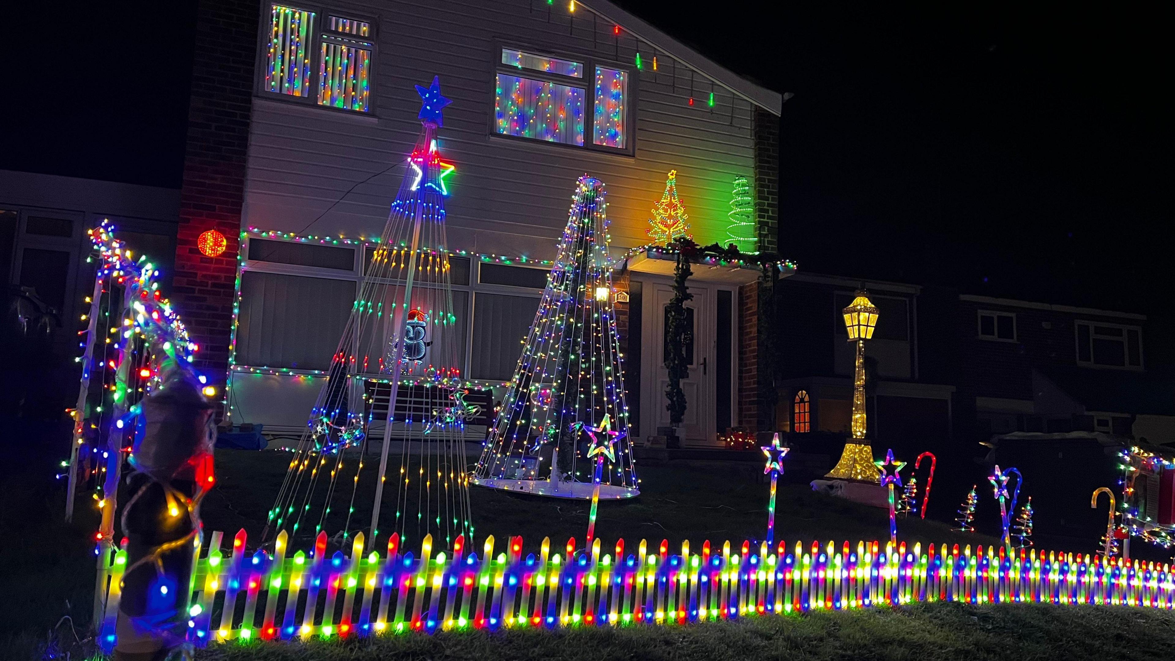 The front garden of a house is filled with lights. Illuminated stars on sticks and poles are made to look like Christmas trees by using strings of lights. A multicoloured, illuminated fence stands at the front of the property.