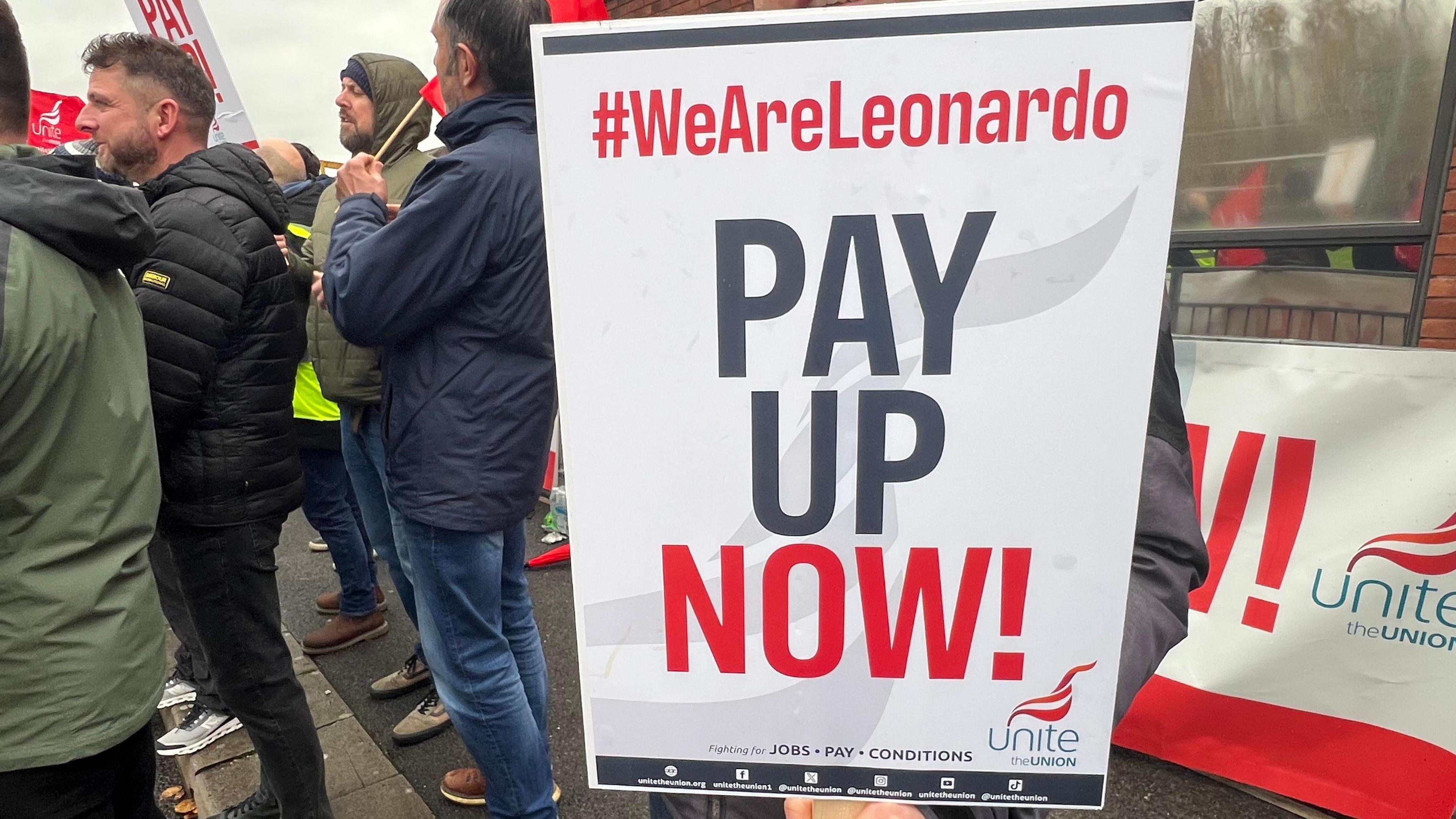 A placard says We Are Leonardo - Pay Up Now. You can see striking union members in the background.