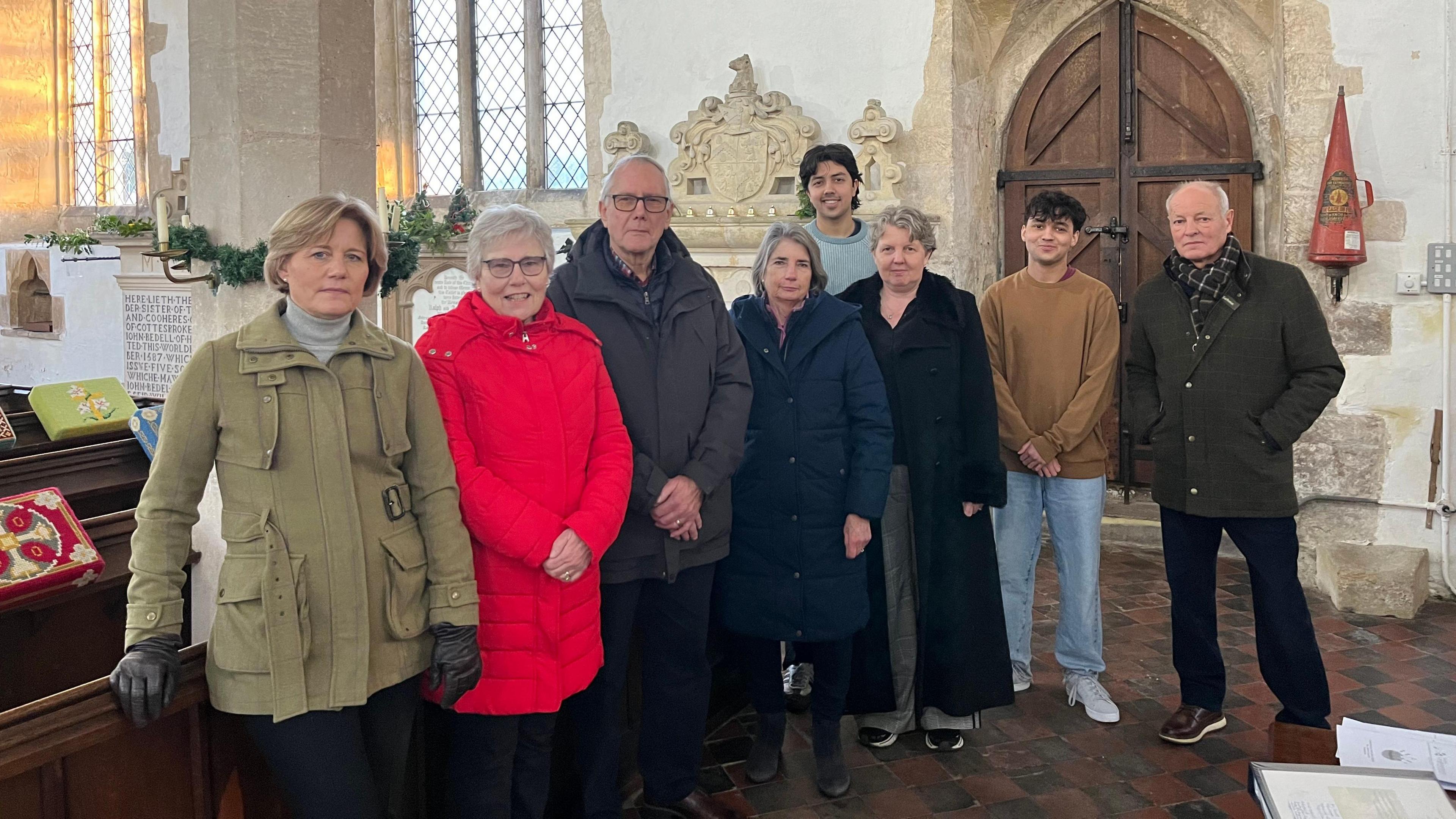 Eight people stand in a line in a church with an entry door behind them, they are members of the congregation. They are all wearing coats and have unsmiling expressions. 