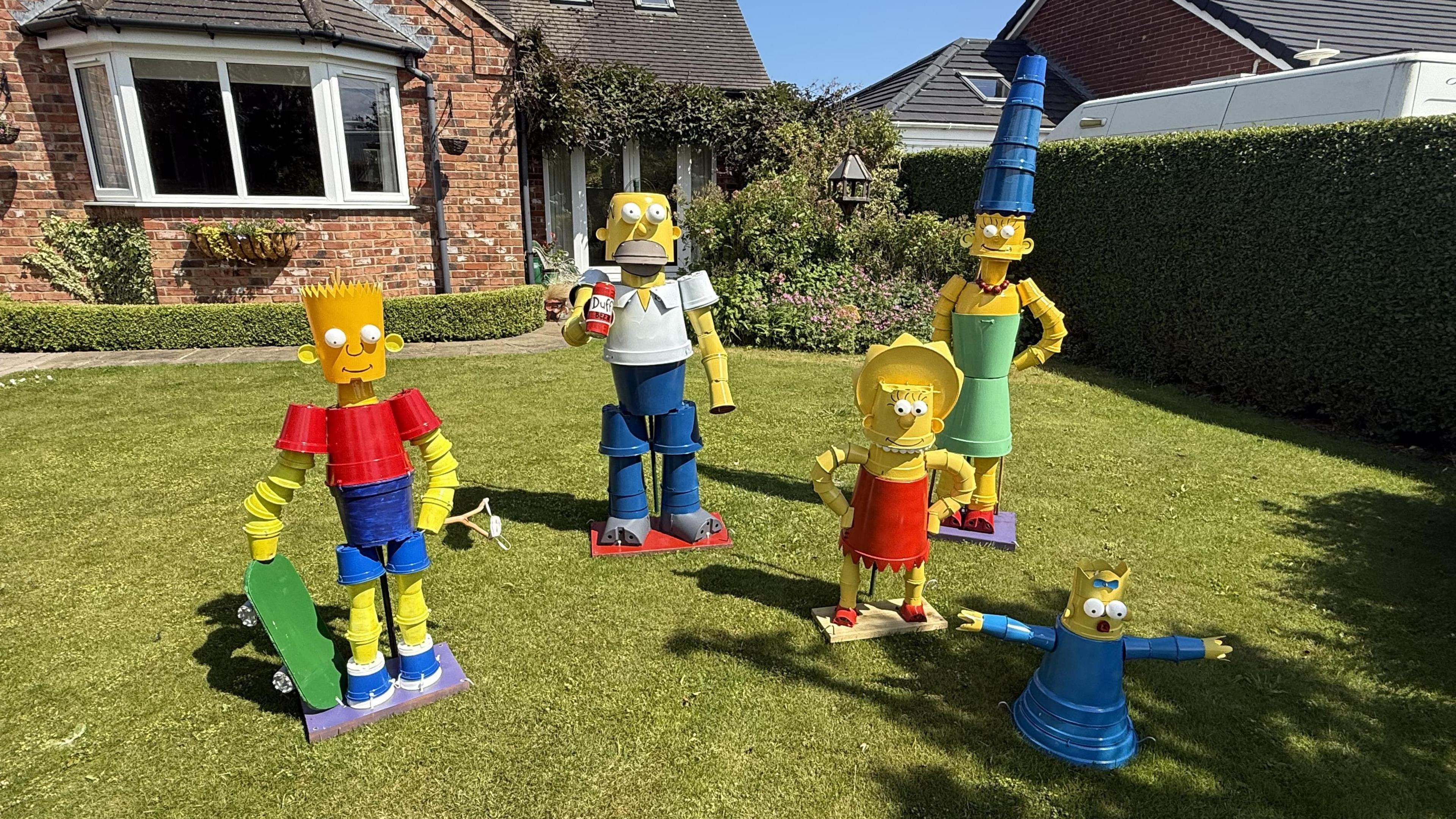 Five statues depicting Bart, Homer, Marge, Maggie and Lisa from the TV show The Simpsons stand on a green lawn. A brick house is in the background.
