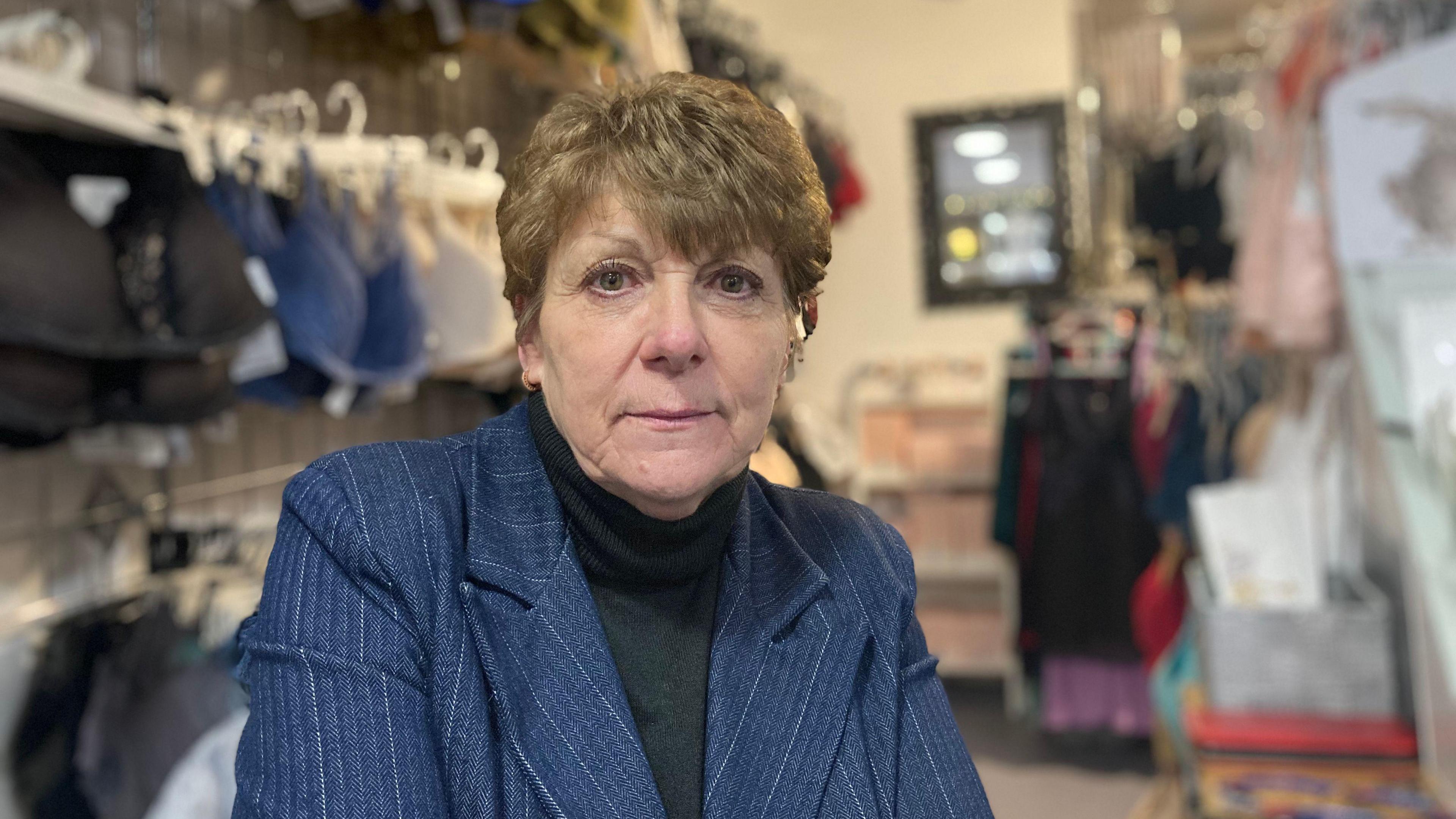 A woman in a black roll neck and navy blazer stands in her shop with women's clothes in the background. 