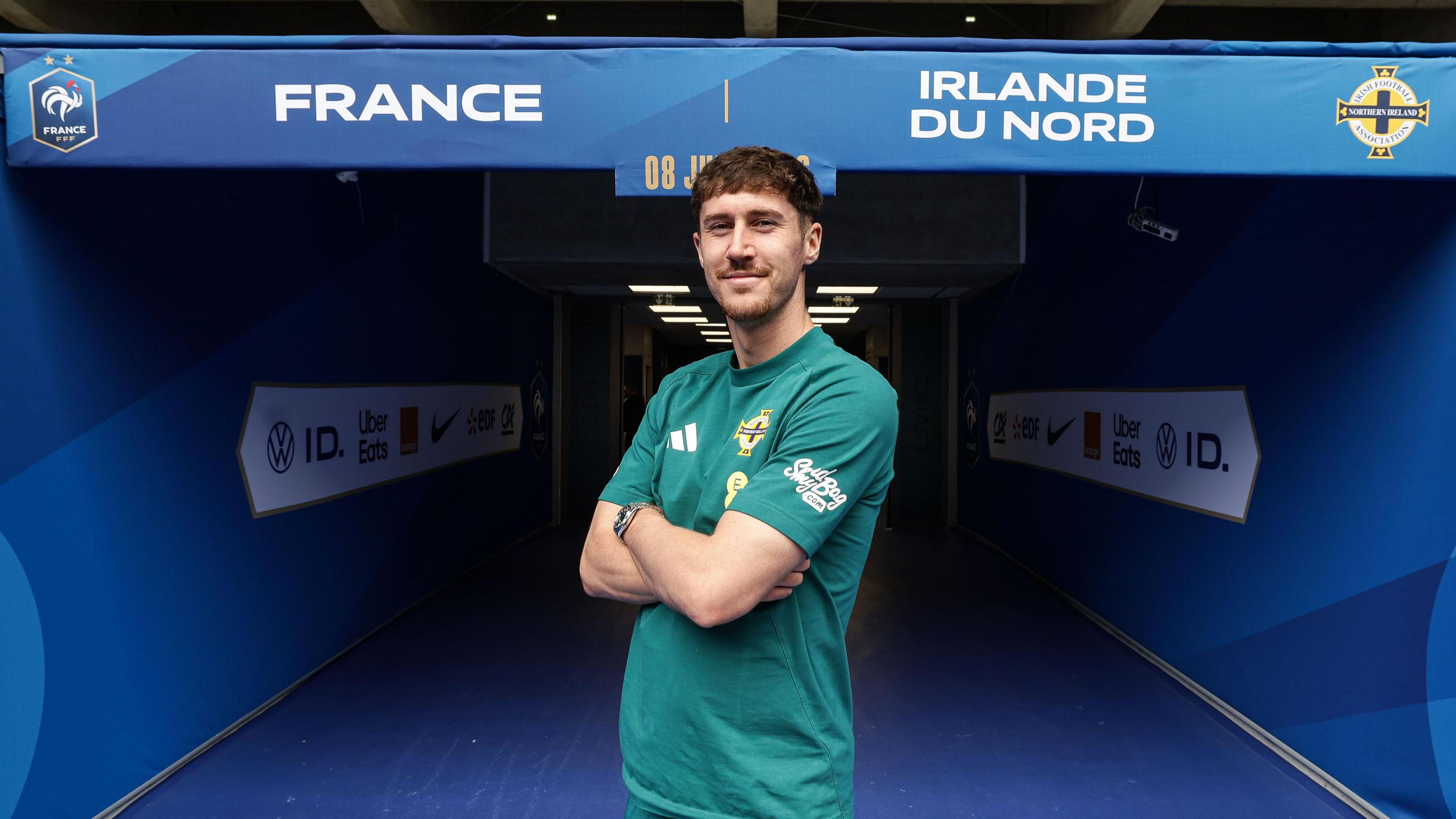 Trai Hume stands in front of the tunnel at the Stade Pierre Mauroy with his arms crossed and facing the camera