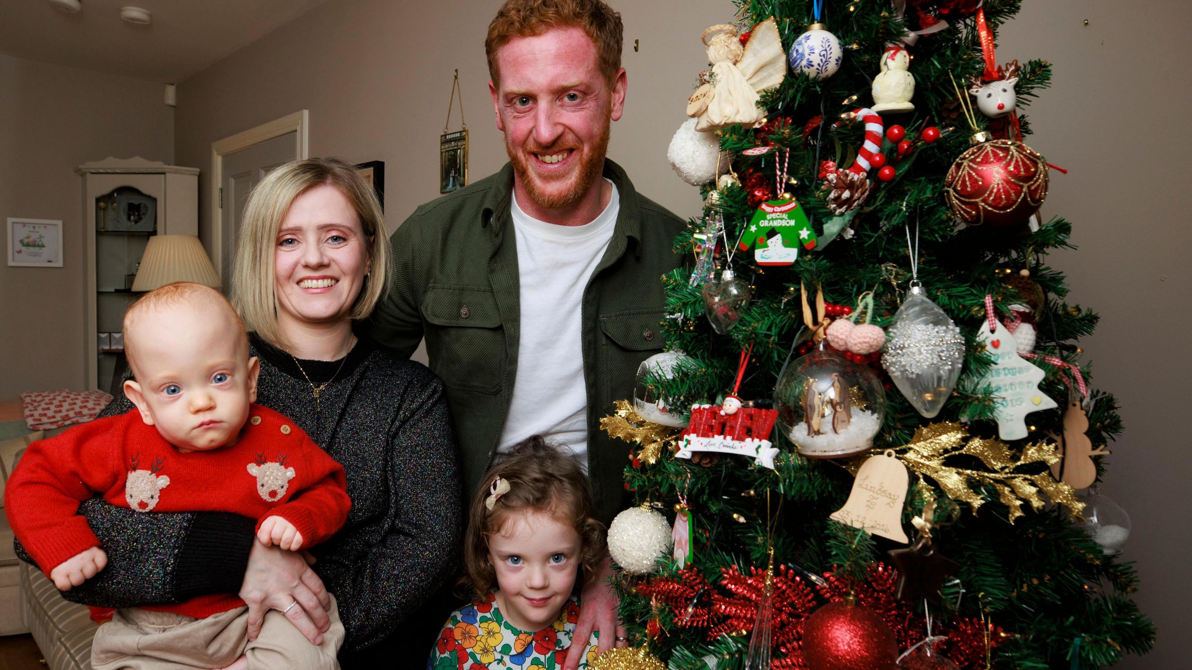 Lindsay Ace, Matthew Ace, four-year-old Aine Ace and Iarla Ace stand beside a christmas tree. Lindsay is smiling and holding Iarla. Matthew Ace has his hand on the shoulder of Aine as they smile.