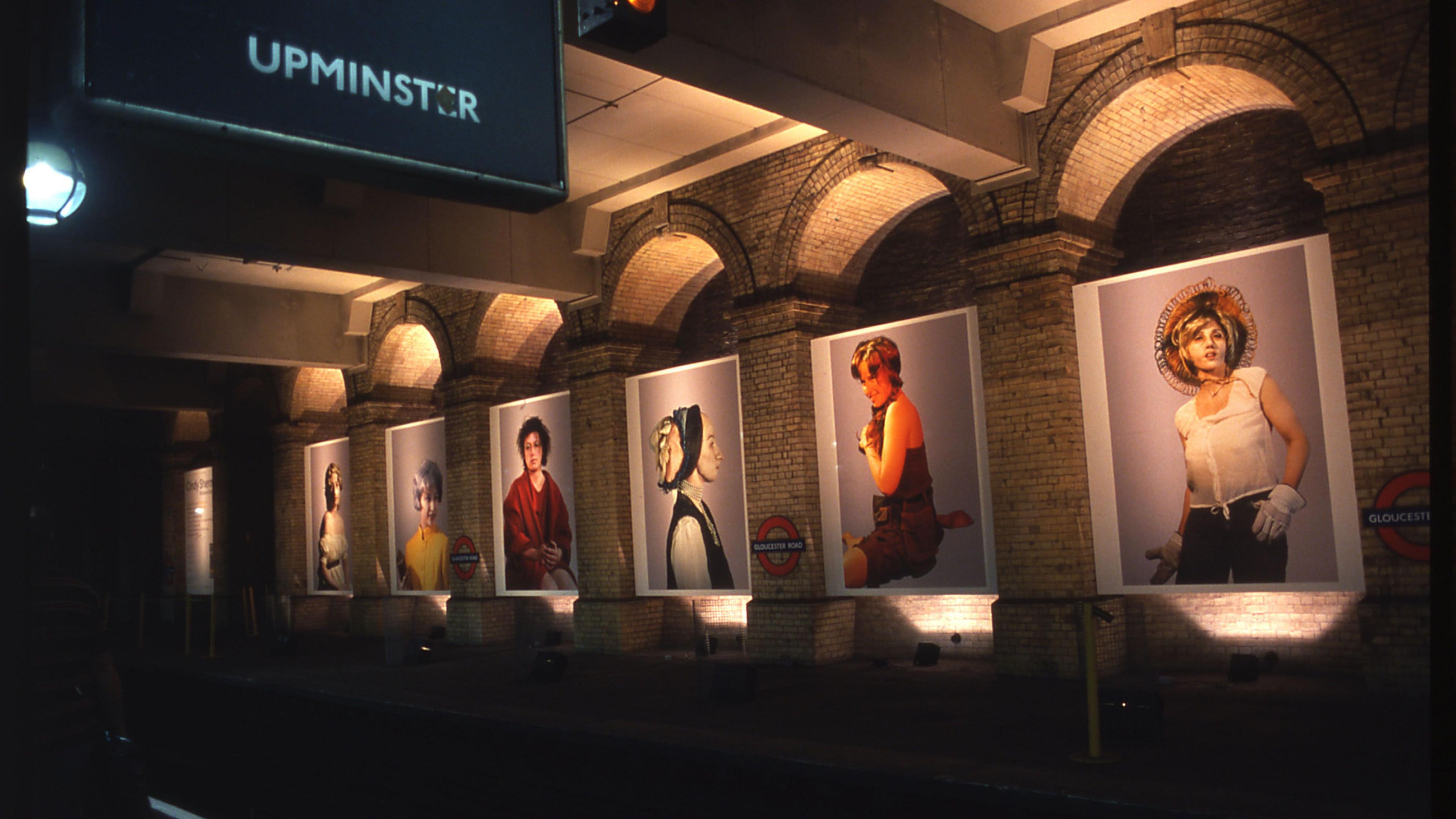 A row of six portraits of women in a row along a Tube platform and lit from below. 