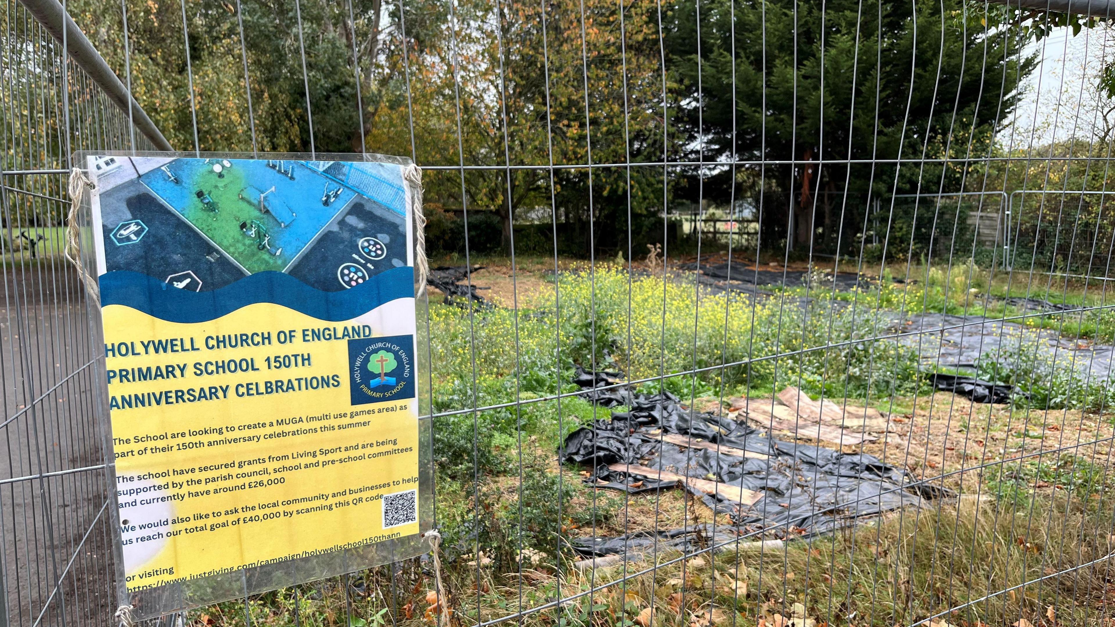 A area of land, roughly 30m square with silver Harris 2m (6ft) high fencing around it, quite a few weeds can be seen and some black membrane sheeting on the ground, there are some large trees in the background.